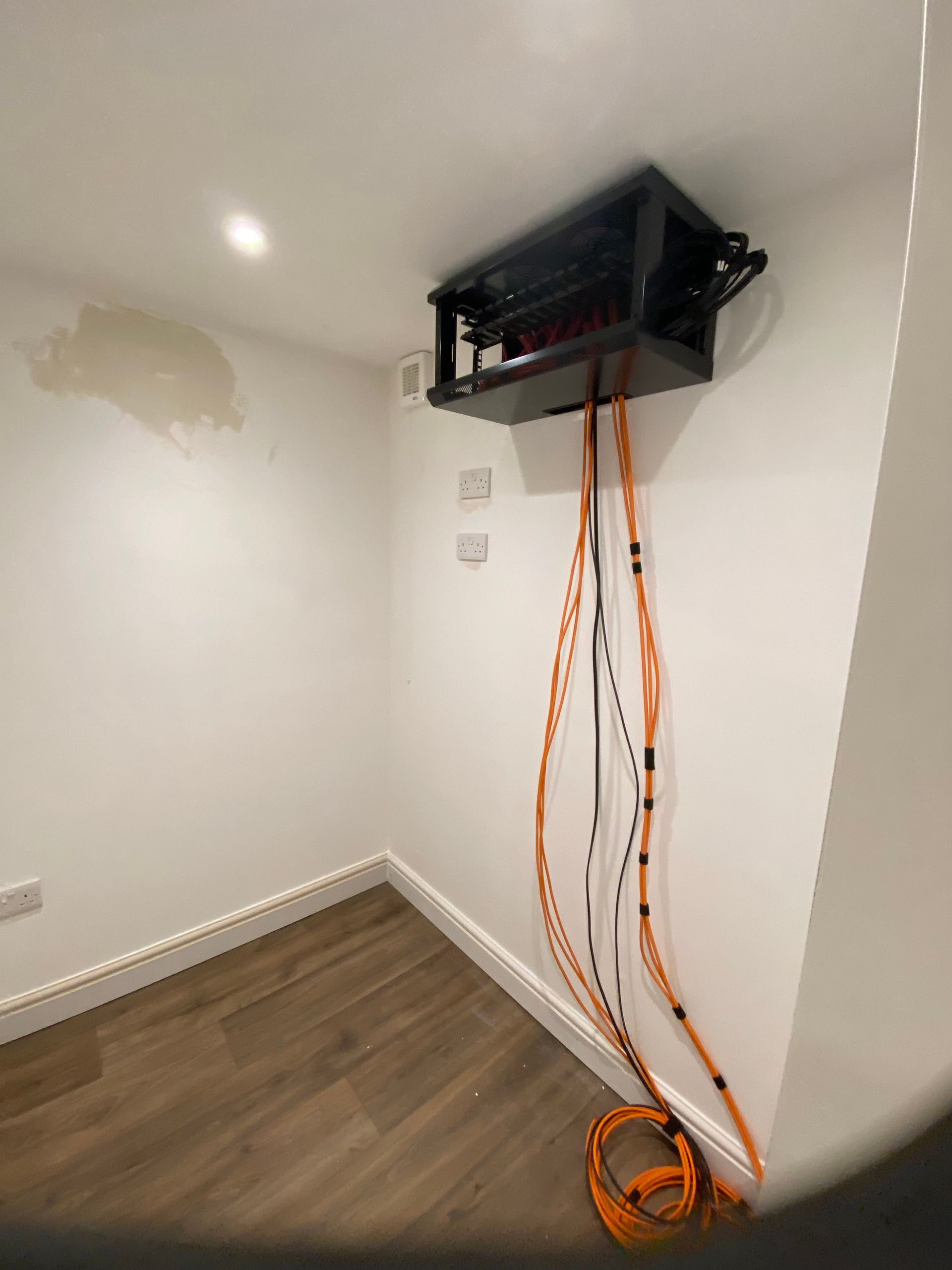 Black patch panel box mounted on a white wall, with orange cables dangling below. Wood flooring.