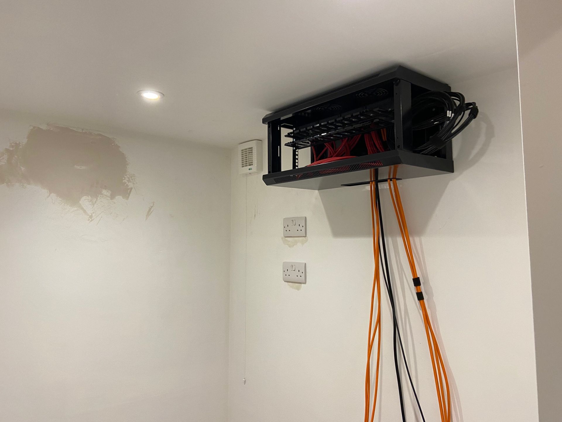 Black patch panel box mounted on a white wall, with orange cables dangling below. Wood flooring.