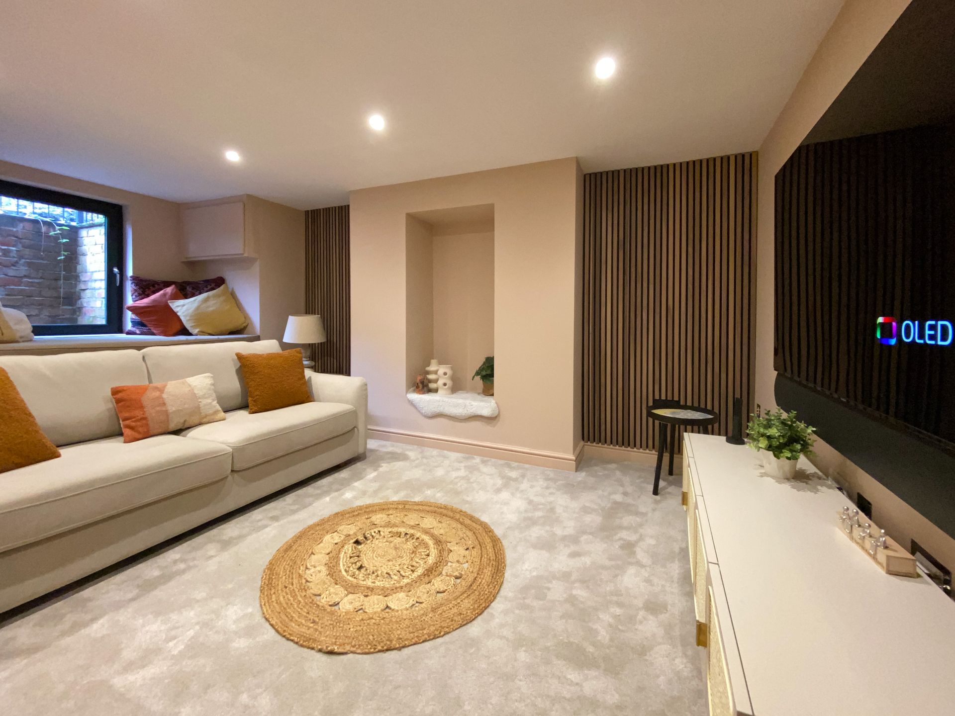 Cozy living room with beige sofa, wooden accent wall, round rug, built-in shelf, and a large television.