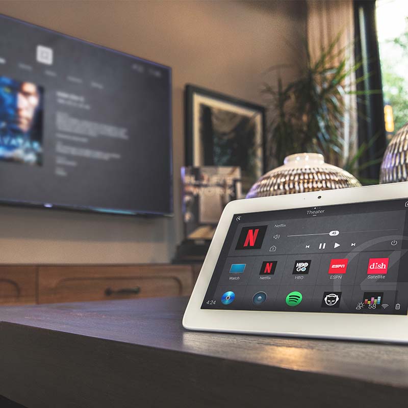Control4 table top touch screen on a table in a room with a large TV in background displaying Control4 user interface