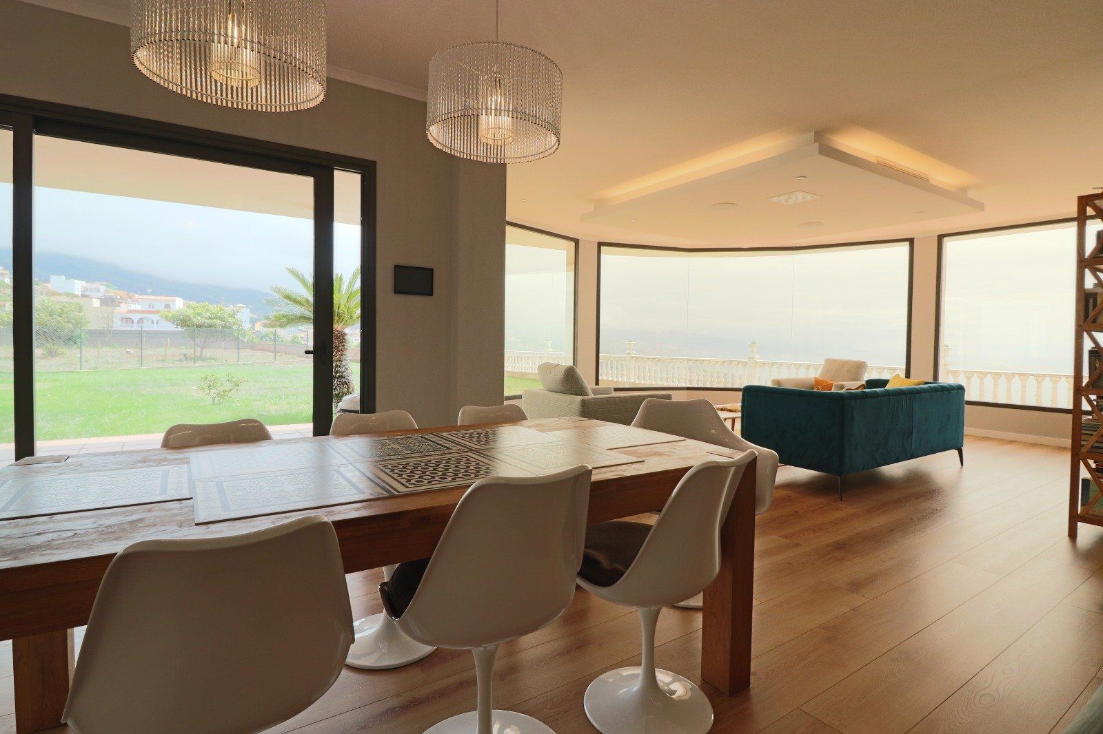 Dining room with a long wooden table, white chairs, and panoramic windows overlooking a green lawn and distant mountains.