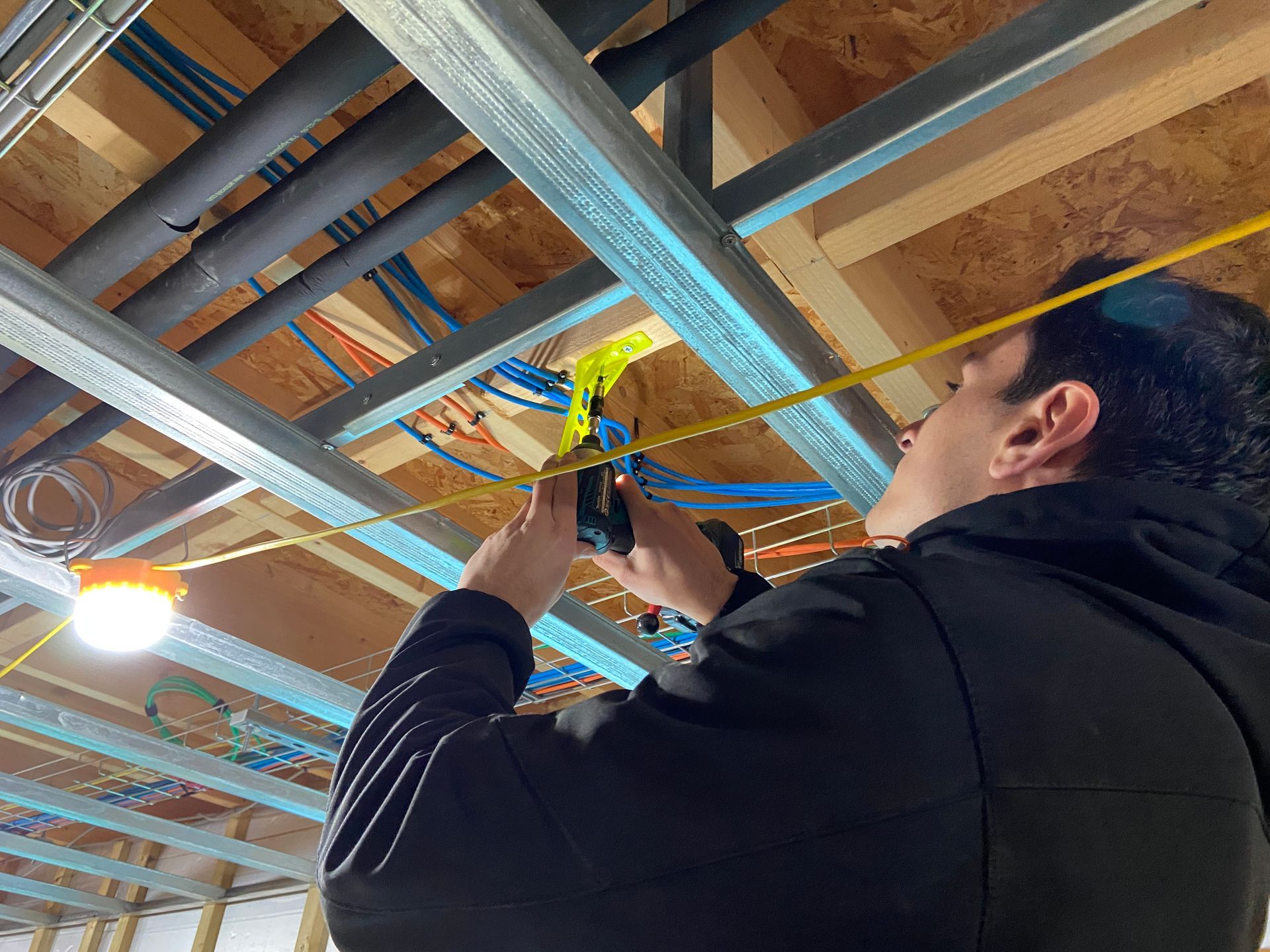 Worker in a black jacket using a power tool to install wiring in a wooden ceiling framework