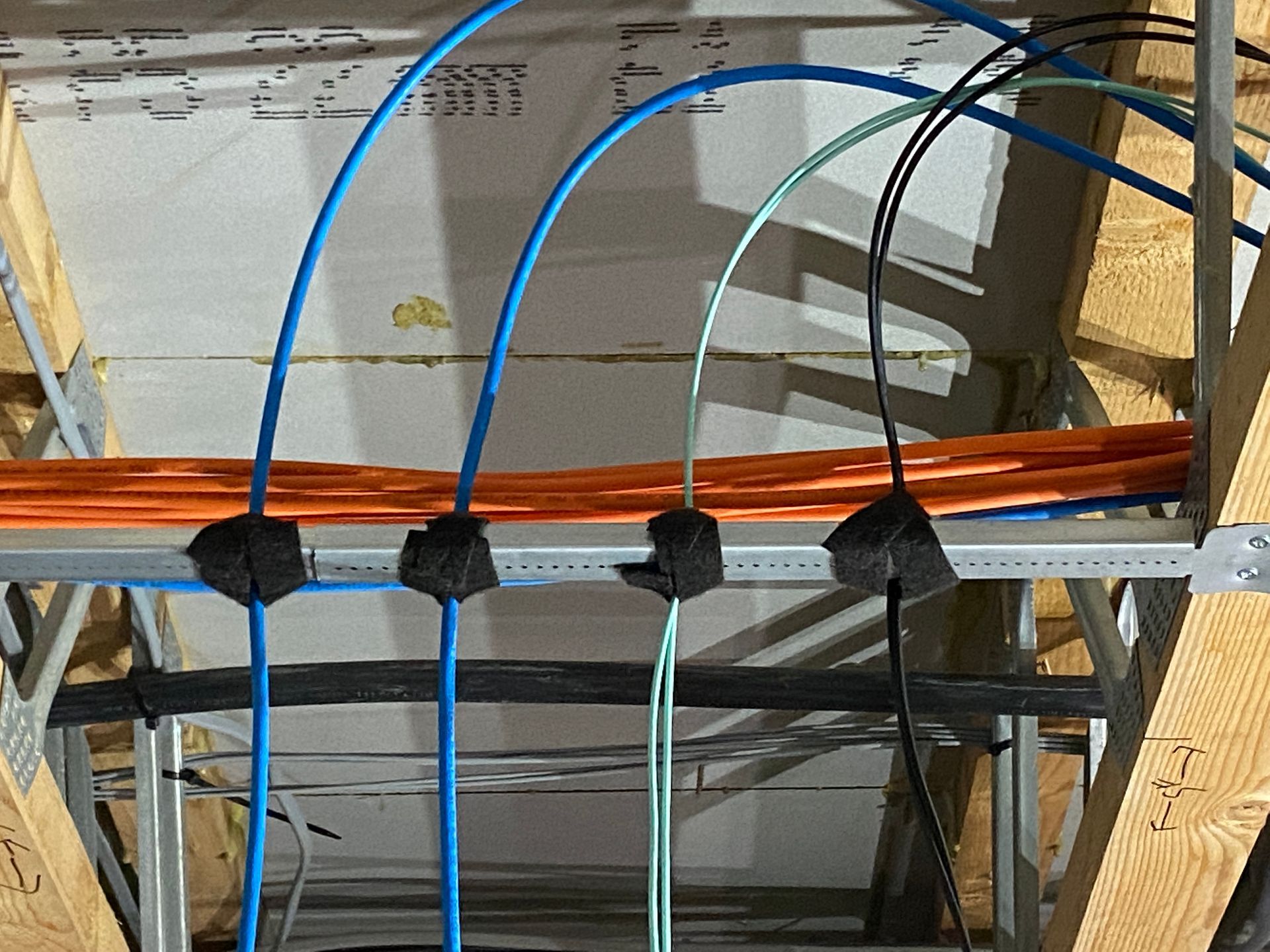 Wires and cables of various colors, including blue, orange, and black, are secured to a metal rack in an unfinished ceiling.