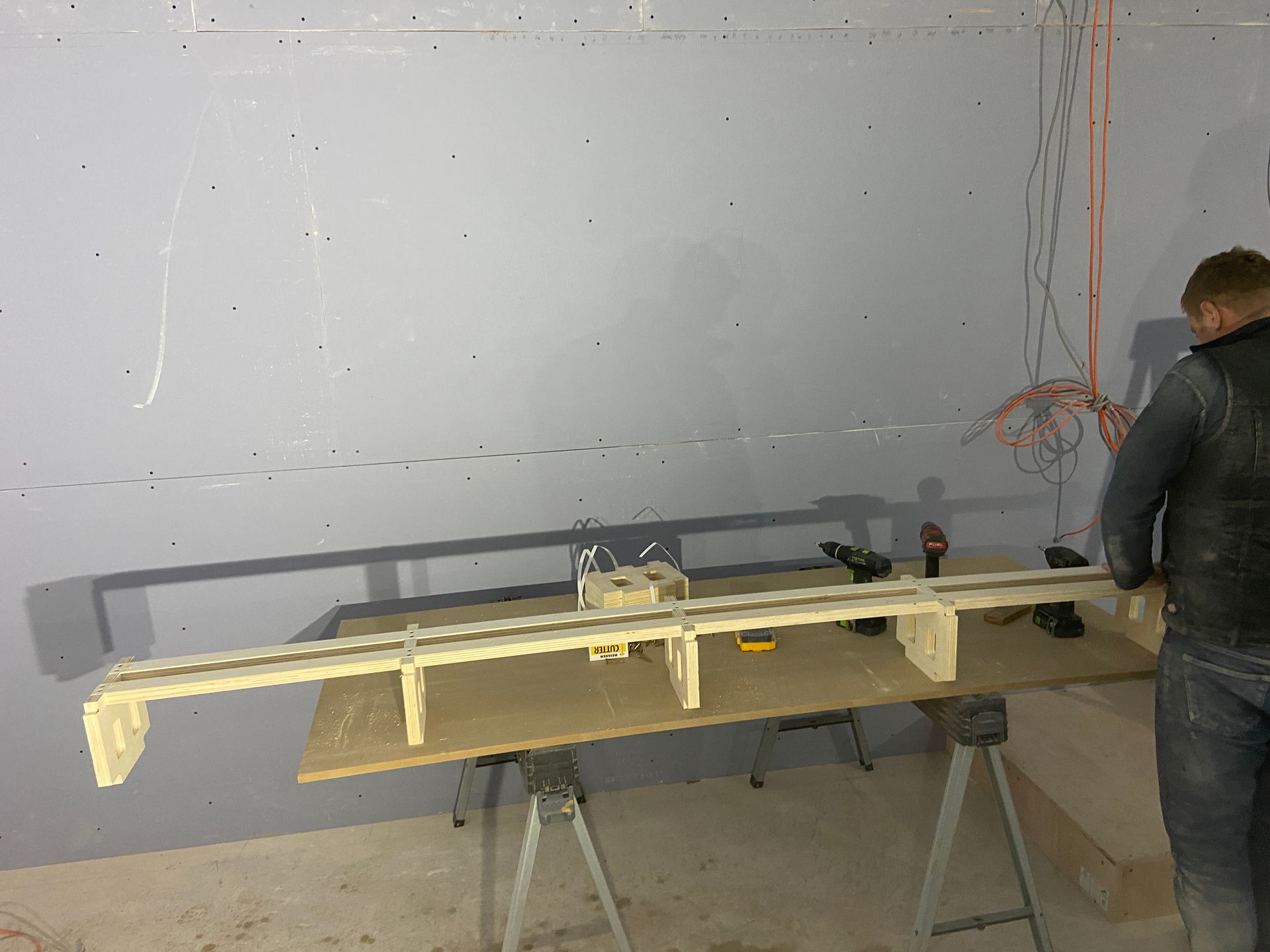 Person works on wooden construction on a table in a room. Electrical wires hang nearby.