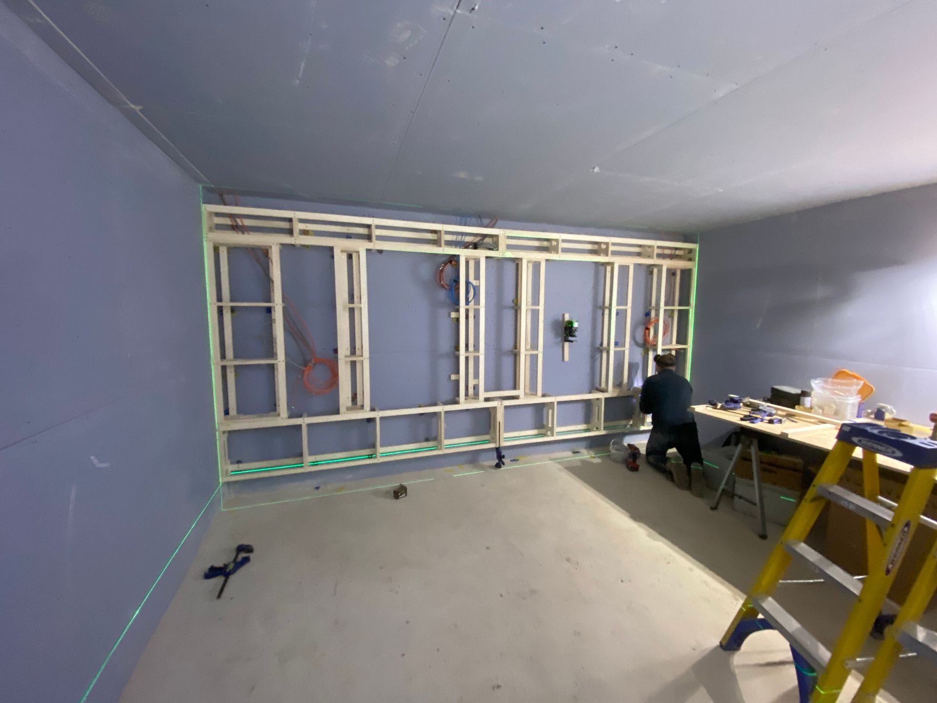 A room under construction; wood frame wall, worker, tools, ladder, and laser level.
