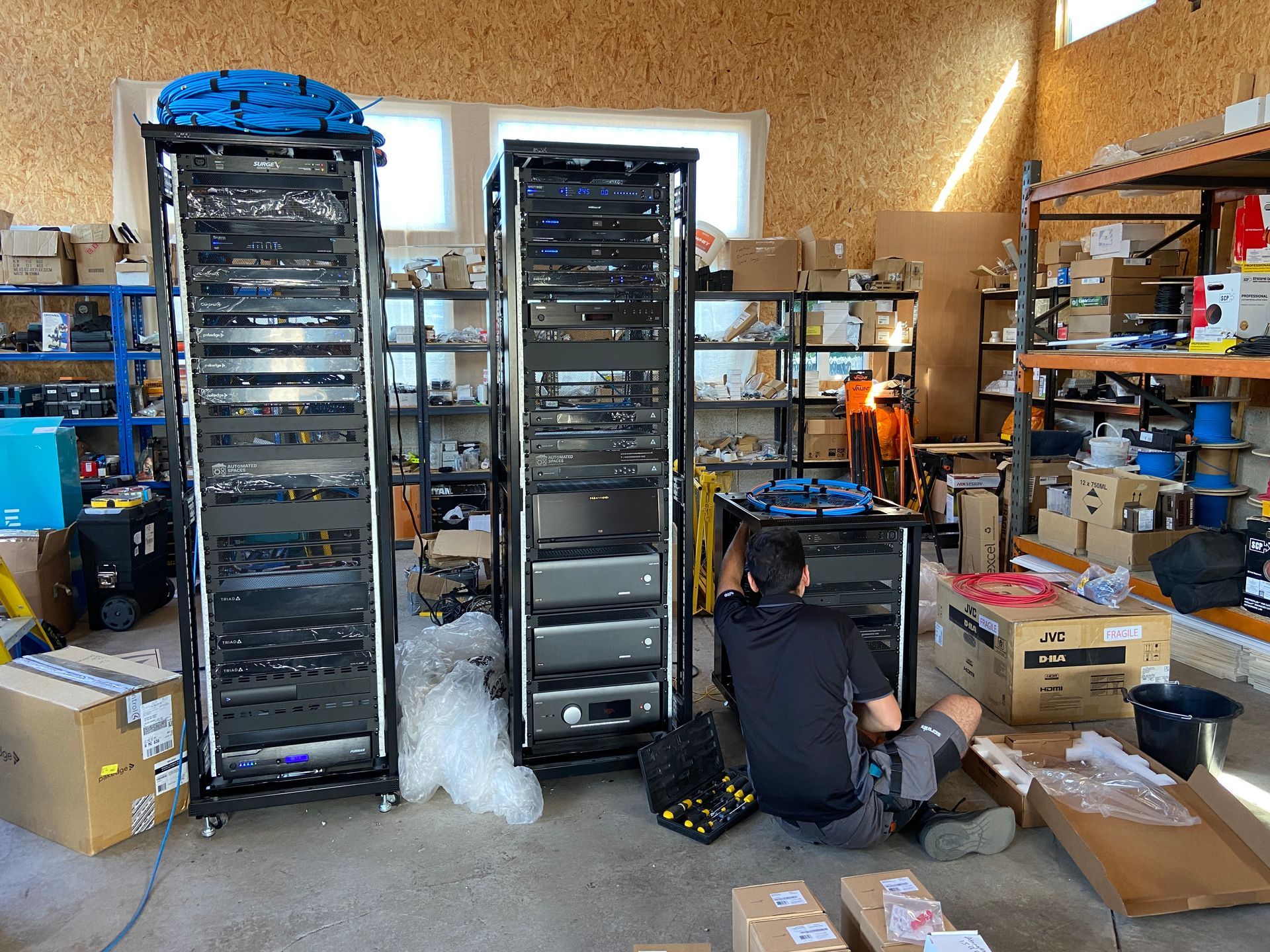 Three black equipment racks in a room. A person works on the middle rack. Boxes and shelves with parts surround.