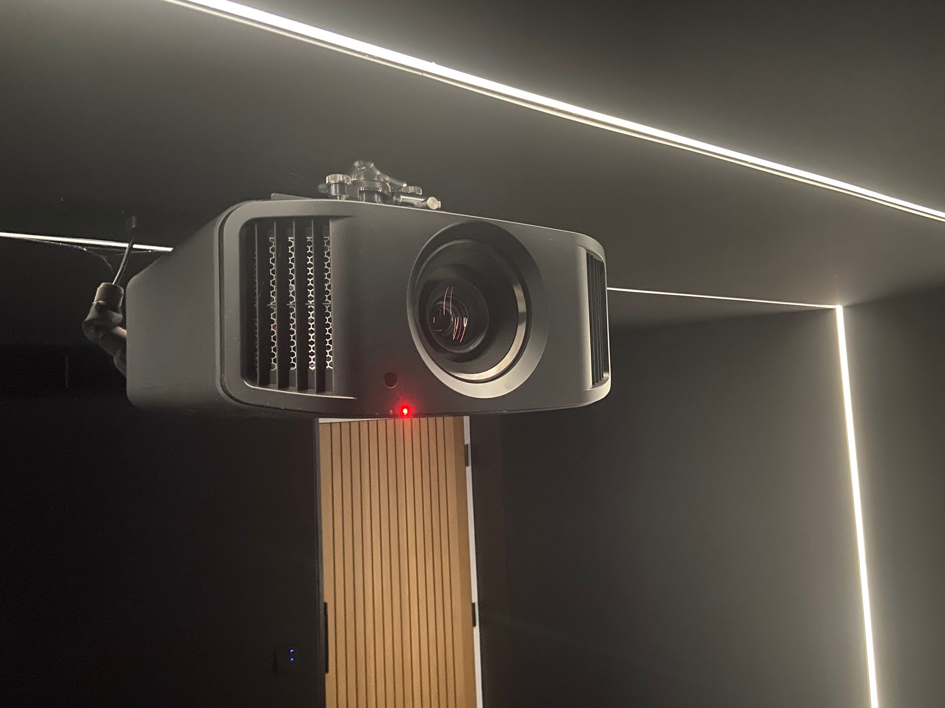 A ceiling-mounted black projector with a red power light, angled towards the viewer, in a dark room with linear lighting.