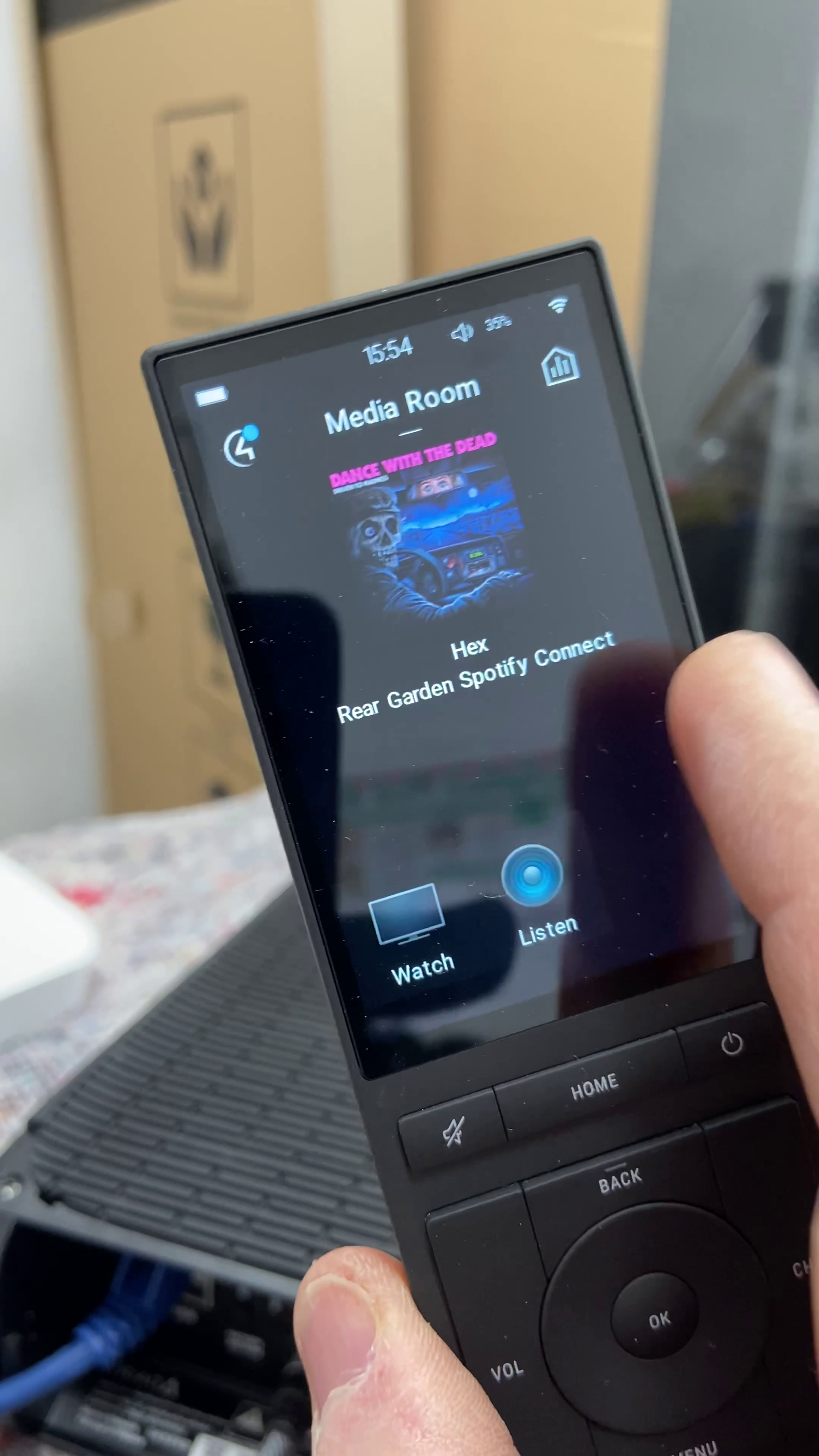 Control4 Neeo remote with the digital music player displaying a song's artwork and controls, near a black device with cables.