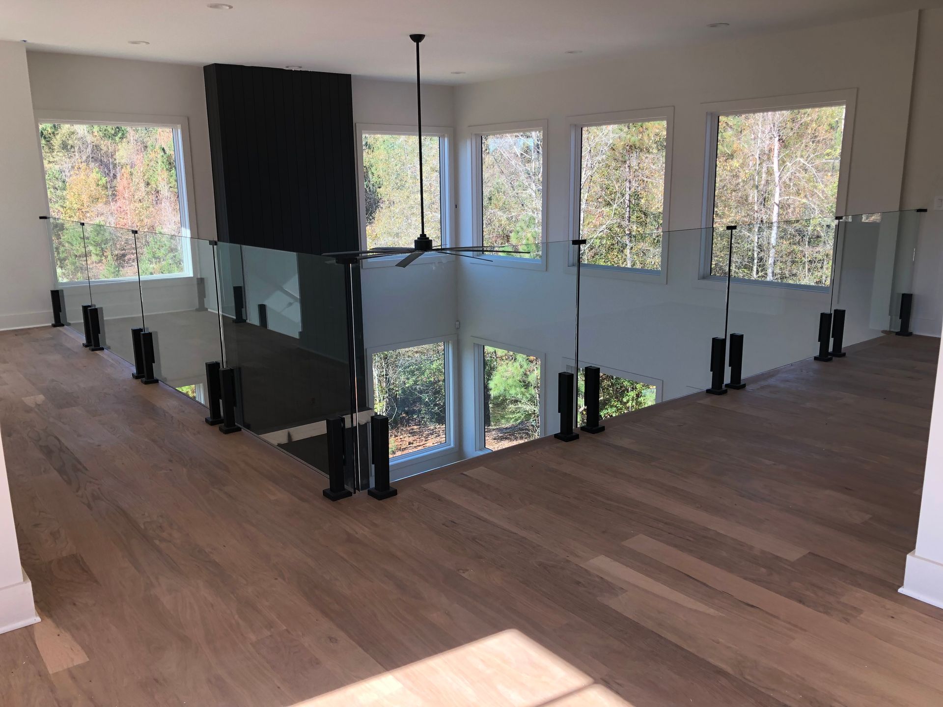 Open loft with wood floors, black railing, and large windows overlooking a bright wooded view.