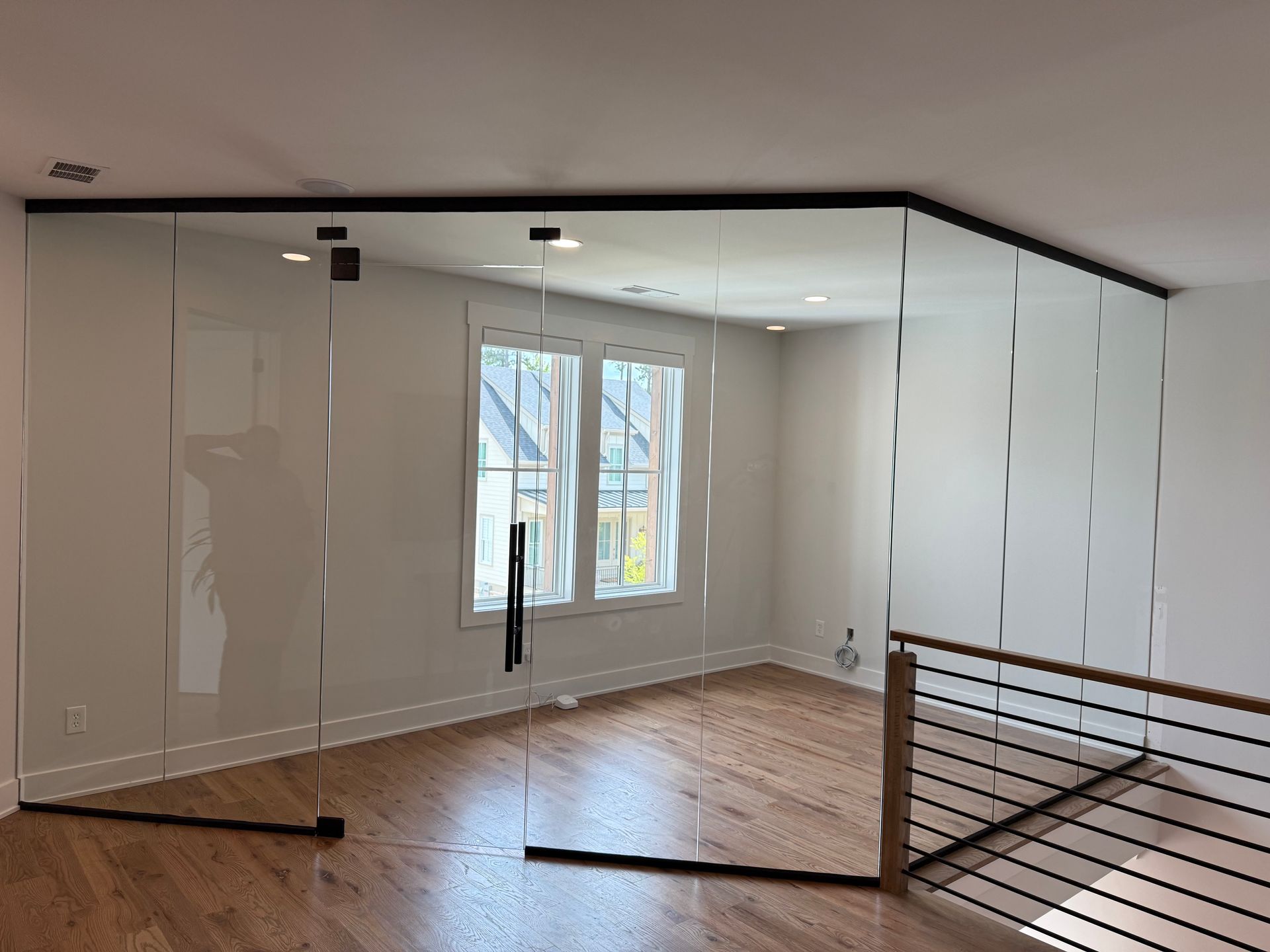 Modern empty room with glossy white walls, wood floor, mirrored closet doors, and a window with black railing.