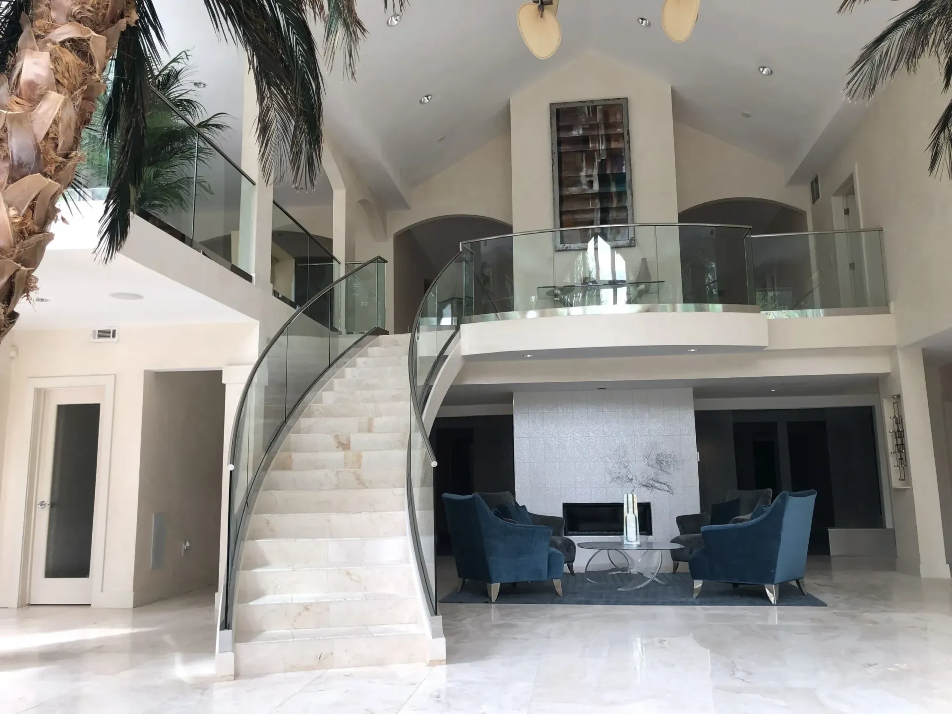 Spacious entryway with marble staircase, glass railings, palm tree, and teal chairs.