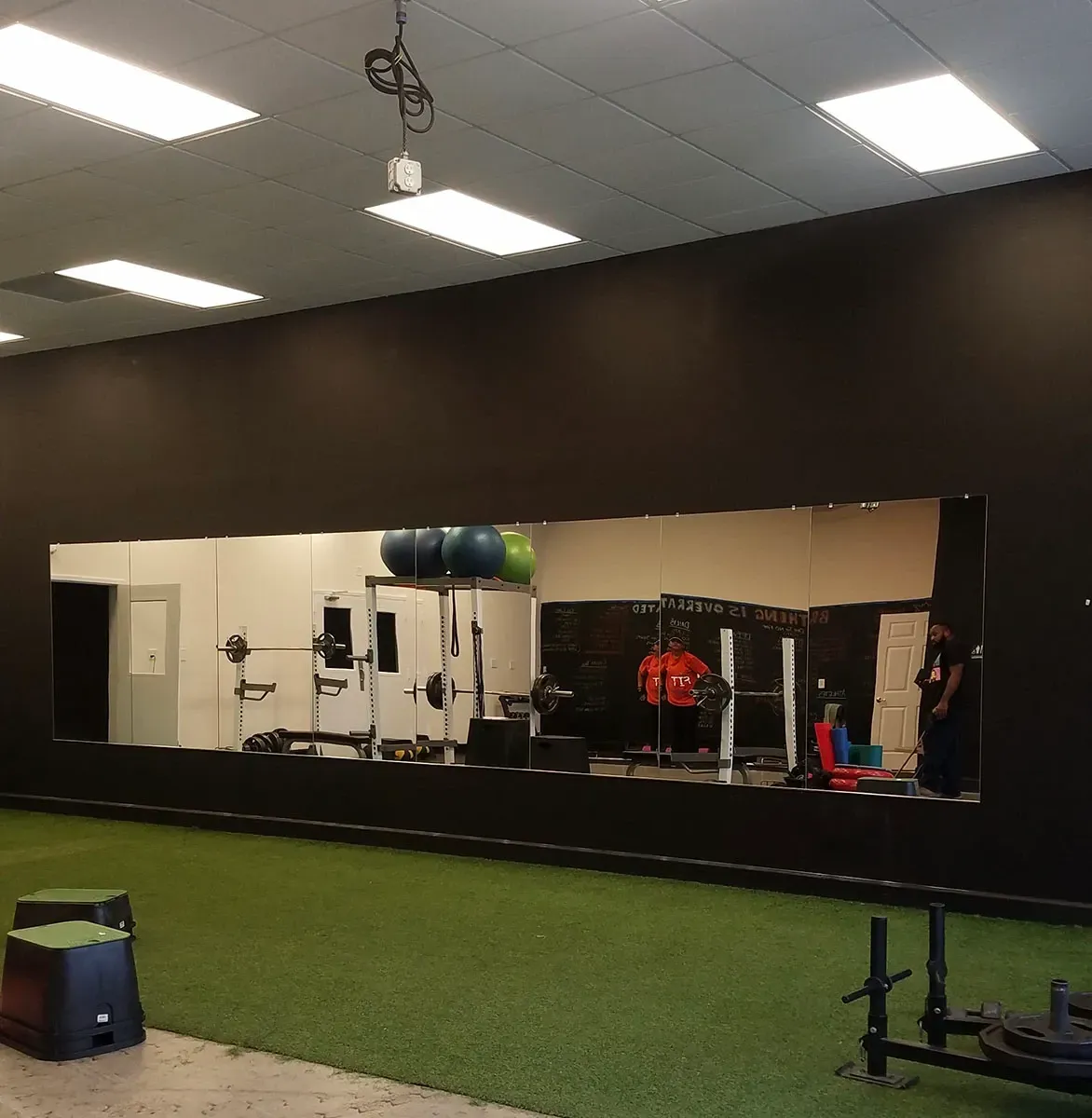 Gym interior with large mirror reflecting workout equipment and people. Green turf floor, black wall.