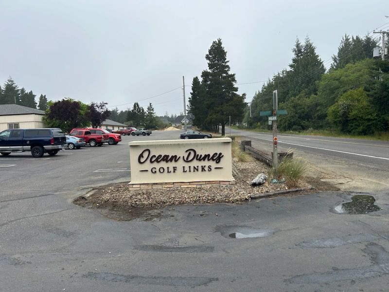Ocean Dunes Golf Links roadside sign with parking lot, trees, and cloudy sky
