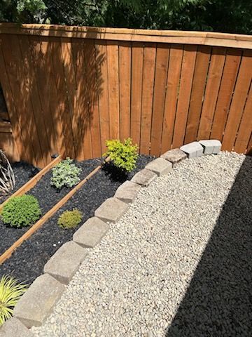 Backyard garden bed with small shrubs, gravel, and a curved stone border beside a wooden fence