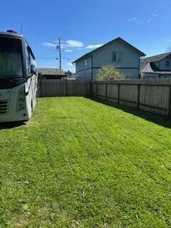 Grassy backyard beside a white bus, with a wooden fence and two houses under a blue sky.