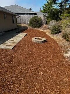 Residential yard with fresh mulch, a circular fire pit, and a house beside a wooden fence.