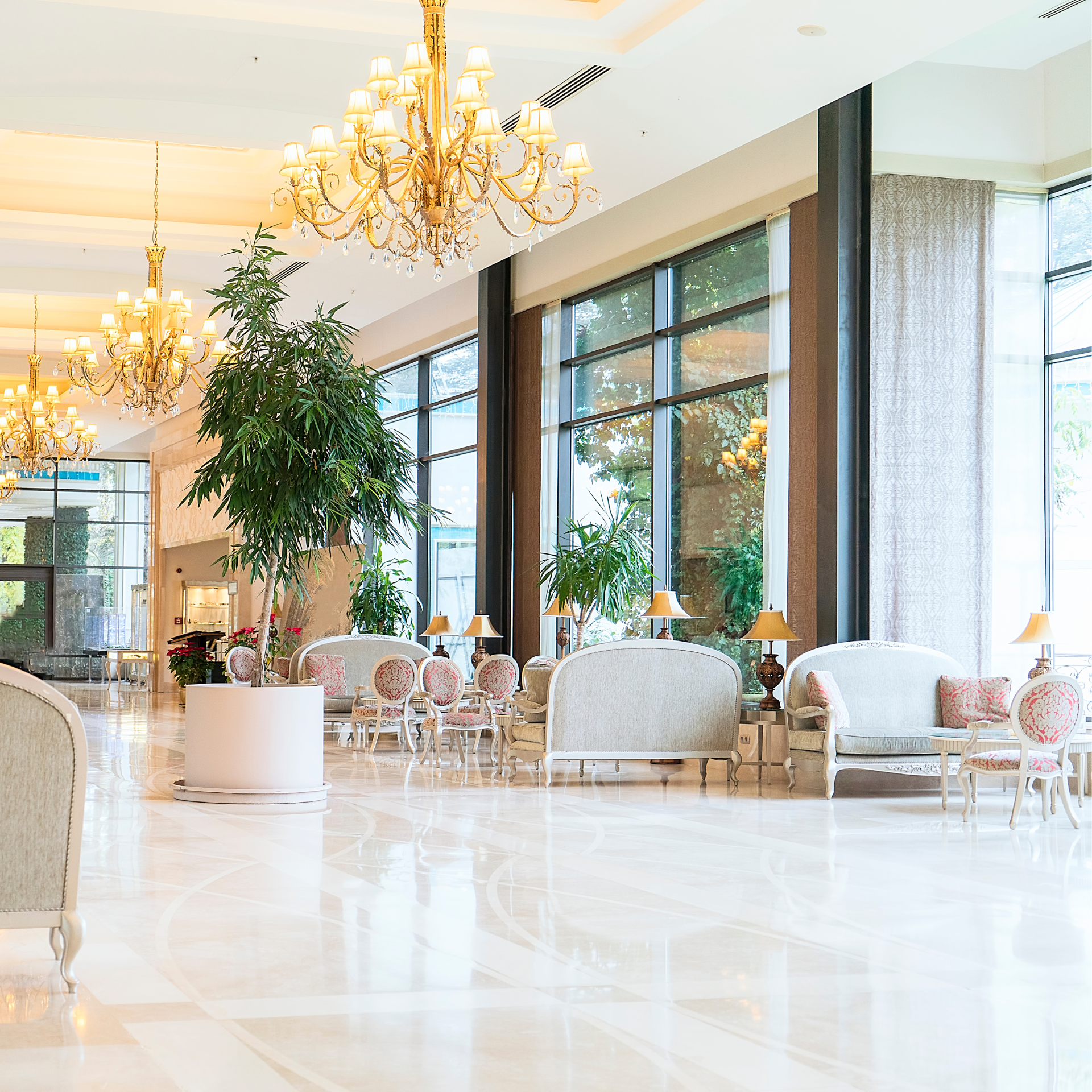 Lobby with chandeliers, couches, large windows, and potted trees. White marble floors.
