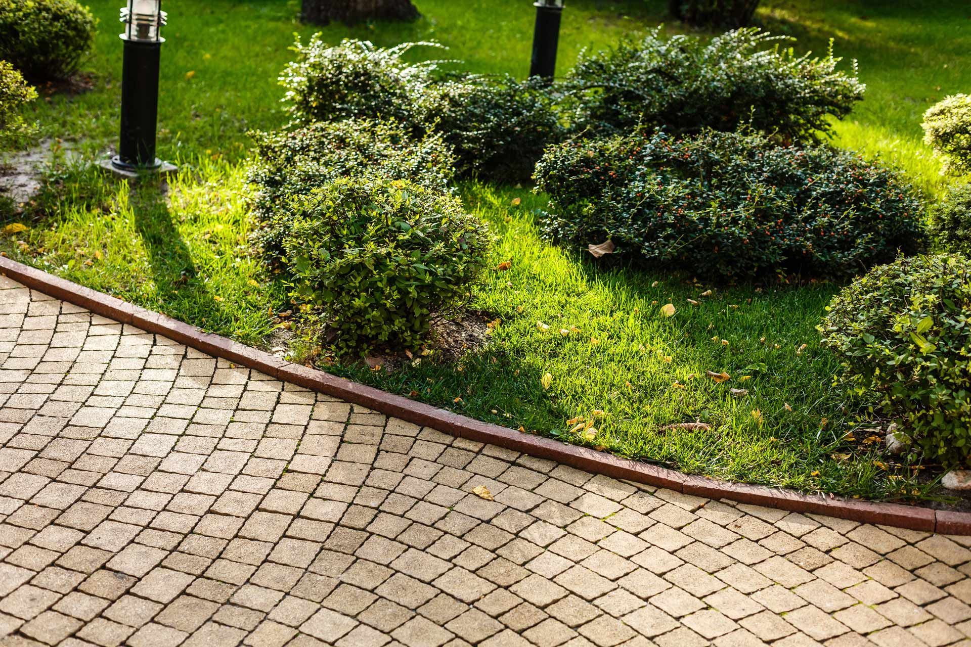 Brick pathway bordering green shrubs and lawn with black light posts.