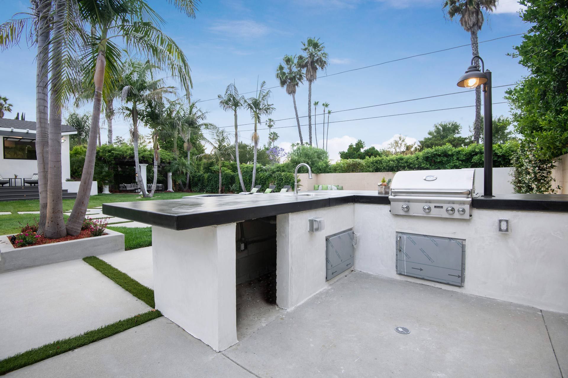 Outdoor kitchen with grill, sink, and concrete countertop, surrounded by palm trees and lawn.