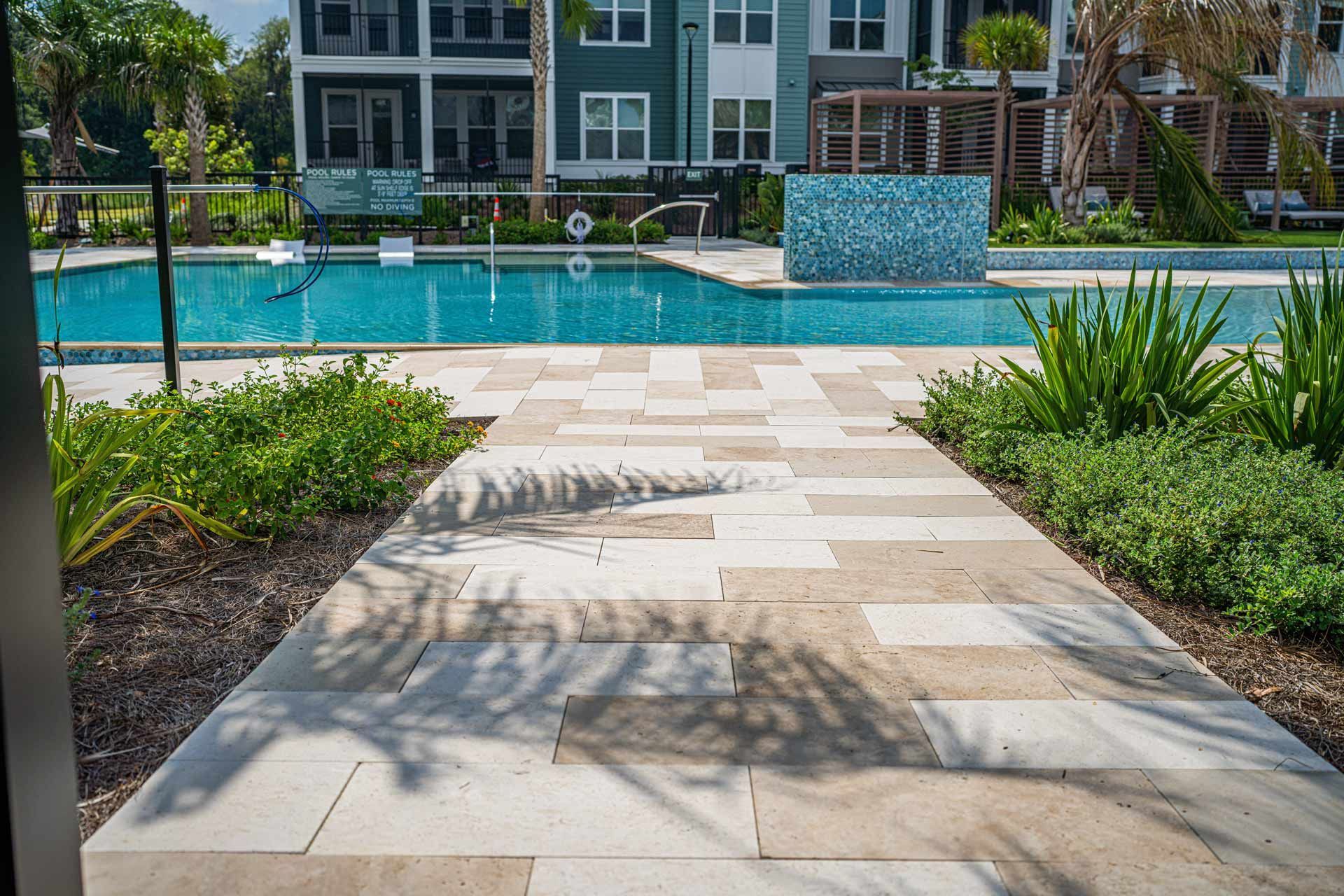 Pathway leads to a swimming pool with modern buildings in the background. Lush greenery lines the path.