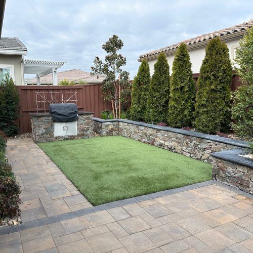 Backyard with stone walls, artificial turf, built-in grill, and evergreen trees.