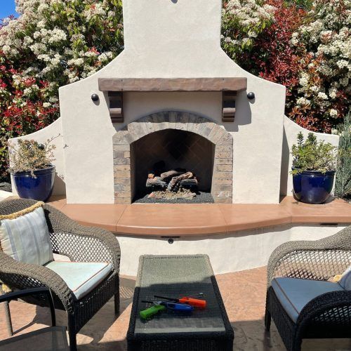 Outdoor fireplace with chairs, table, and blooming bushes in the background.