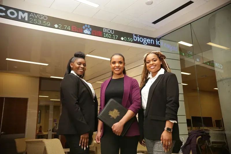 Three women are standing next to each other in front of a stock exchange.
