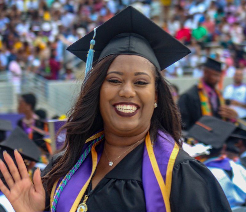 A woman in a graduation cap and gown is smiling and waving at the camera.