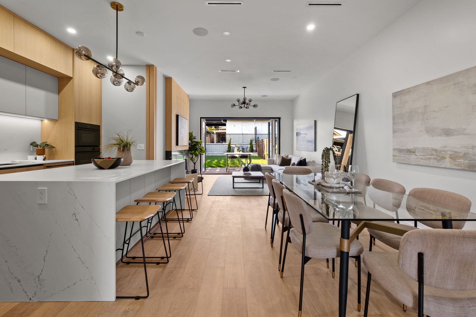 Modern open-plan living space with kitchen, dining area, and outdoor access. Light wood floors, white walls, and contemporary furnishings.