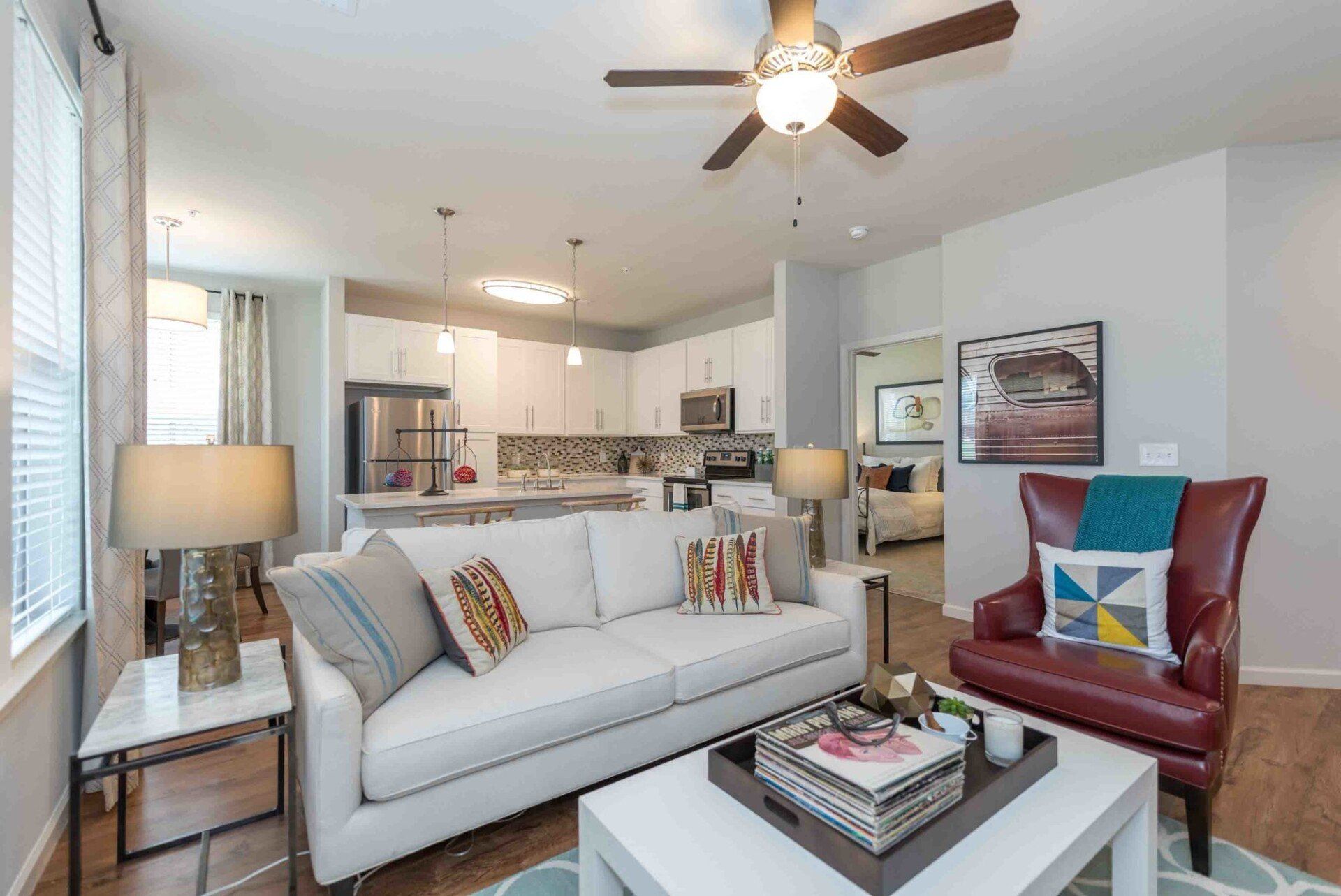 A bright living room features a white sofa, a dark red armchair, a coffee table, and an open-concept kitchen beyond at Silver Collection at Celebrate apartment community in Fredericksburg, VA.