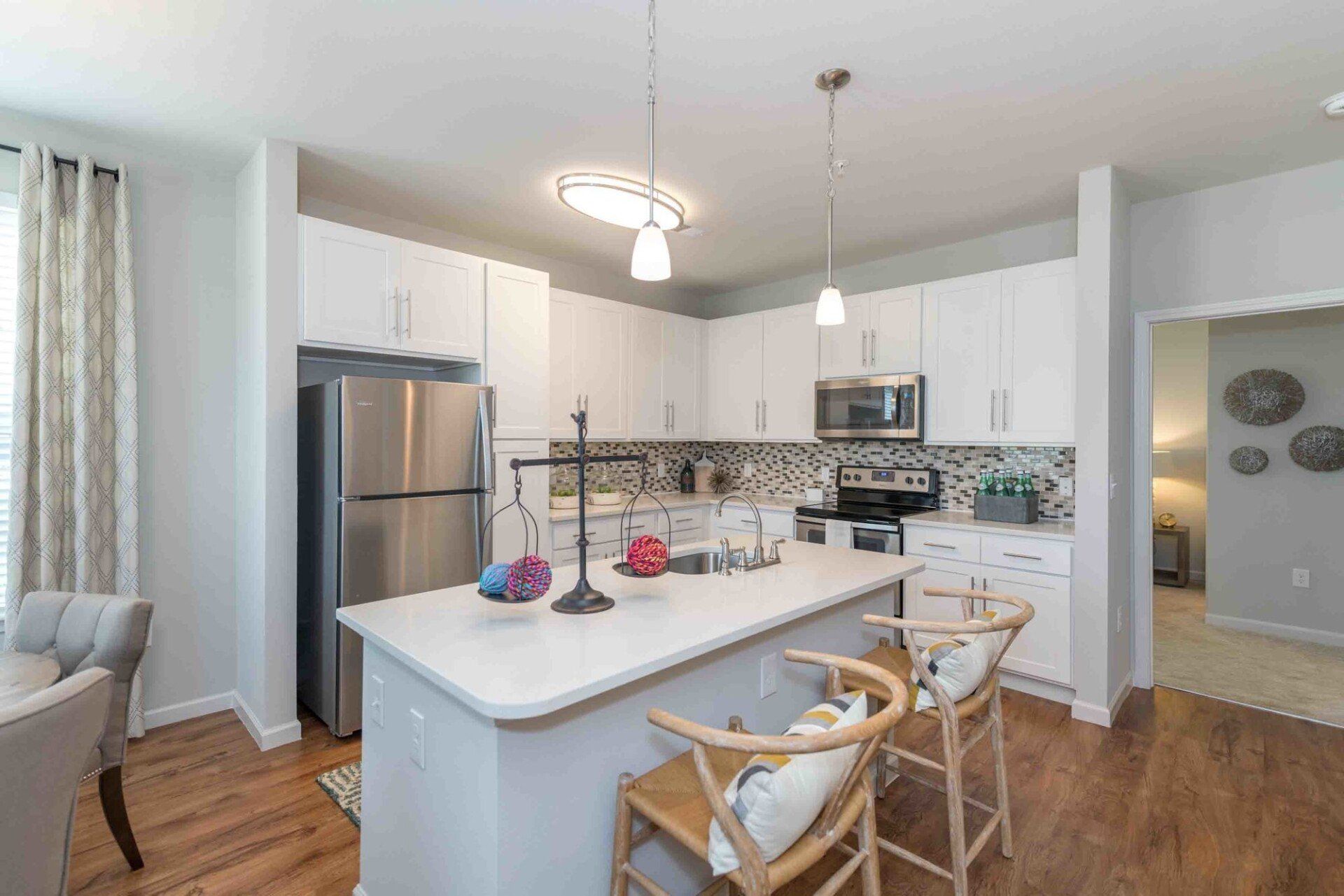 A modern kitchen features white cabinetry, stainless steel appliances, a center island with bar stools, and wood flooring at Silver Collection at Celebrate apartment community in Fredericksburg, VA.