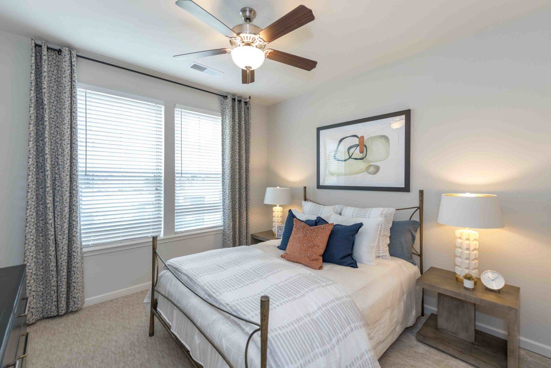 A modern bedroom with a metal-frame bed, patterned bedding, two nightstands with lamps, and gray curtains by a window at Silver Collection at Celebrate apartment community in Fredericksburg, VA.