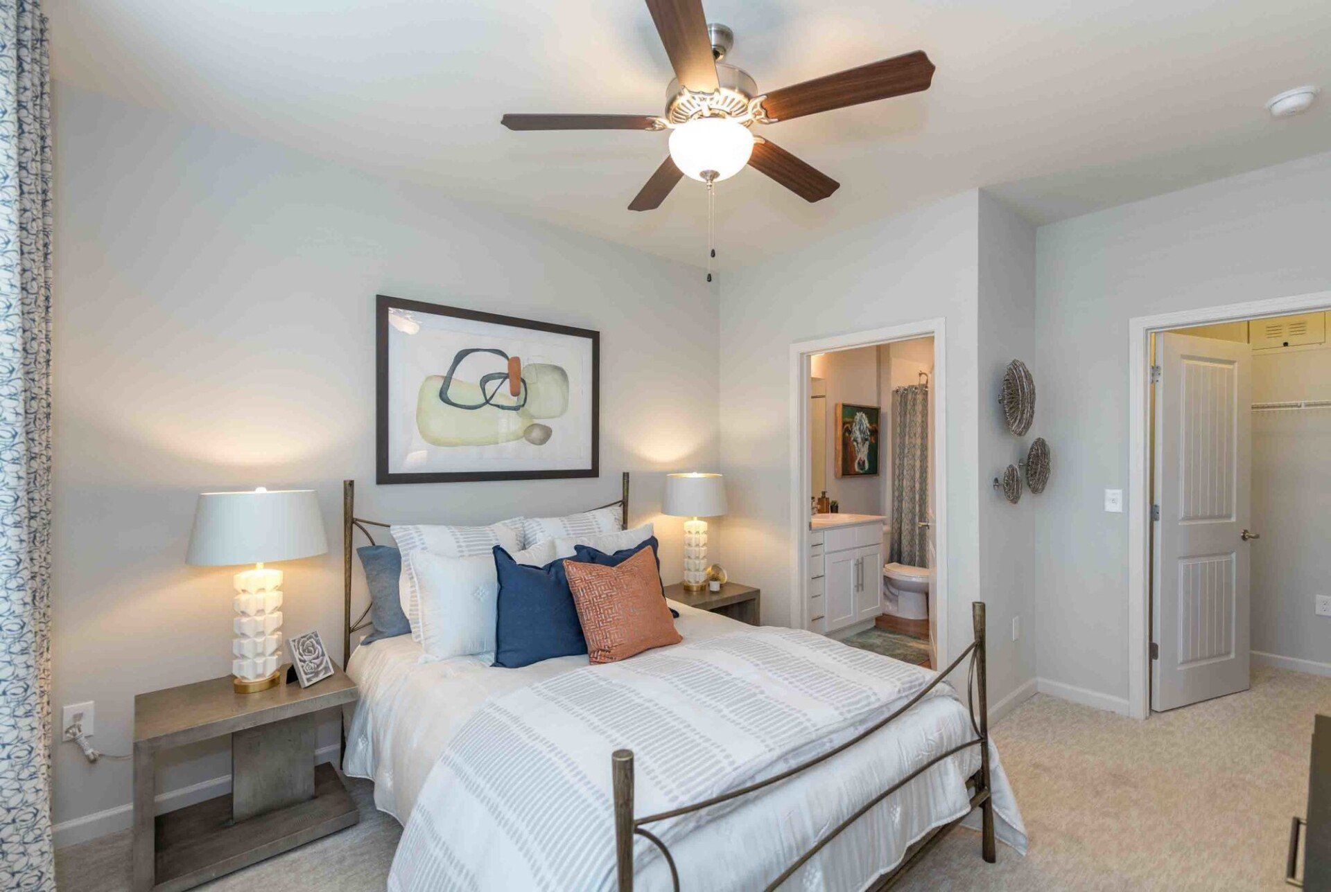 A bedroom with a metal-frame bed, a bedside table with a lamp, a ceiling fan, and an open door to a bathroom at Silver Collection at Celebrate apartment community in Fredericksburg, VA.