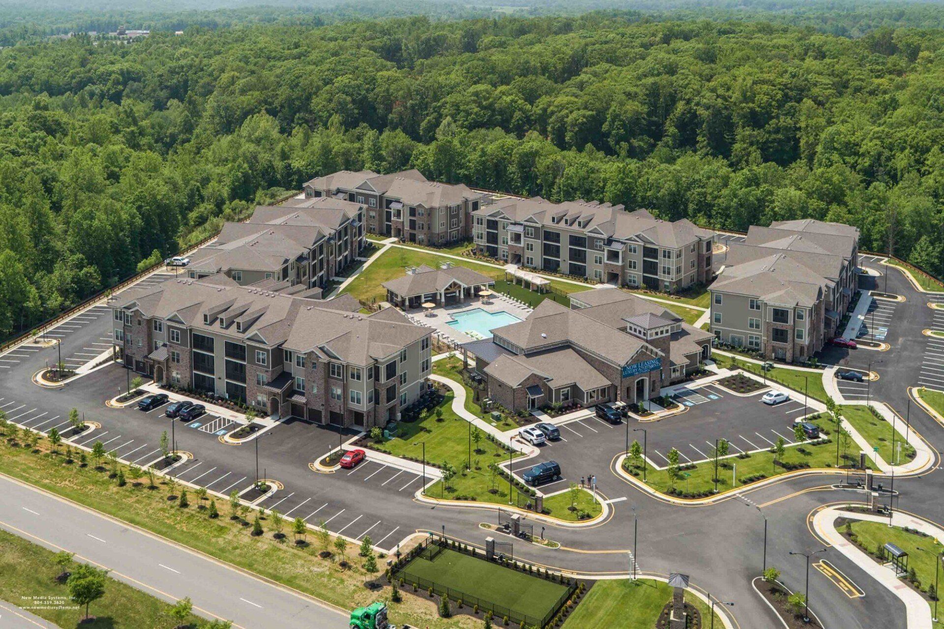 Aerial view of a multi-building apartment complex with a central swimming pool surrounded by woods and parking lots at Silver Collection at Celebrate apartment community in Fredericksburg, VA.