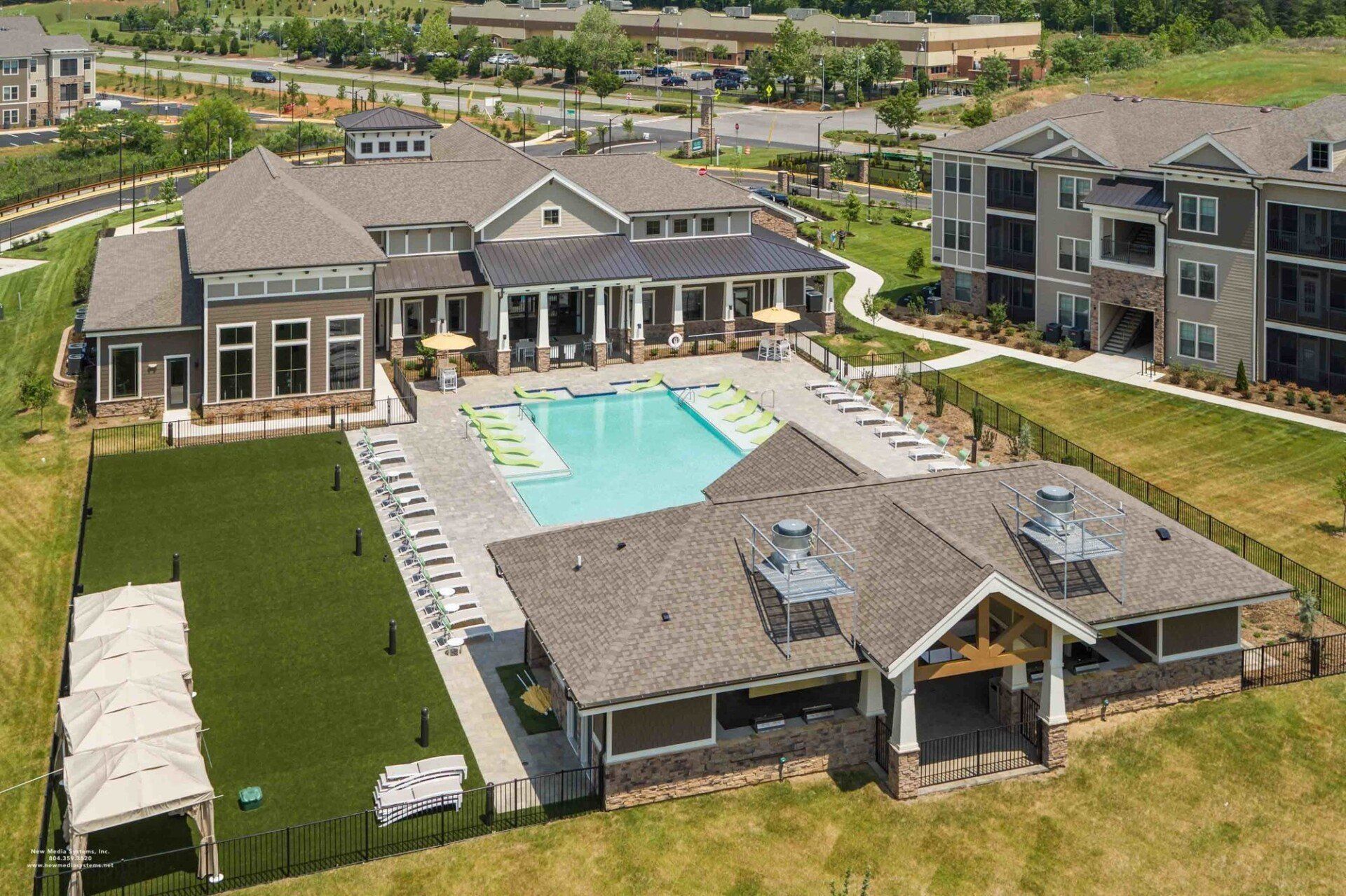 Aerial view of an apartment community with a large outdoor swimming pool, lounge chairs, a clubhouse, and green lawn at Silver Collection at Celebrate apartment community in Fredericksburg, VA.