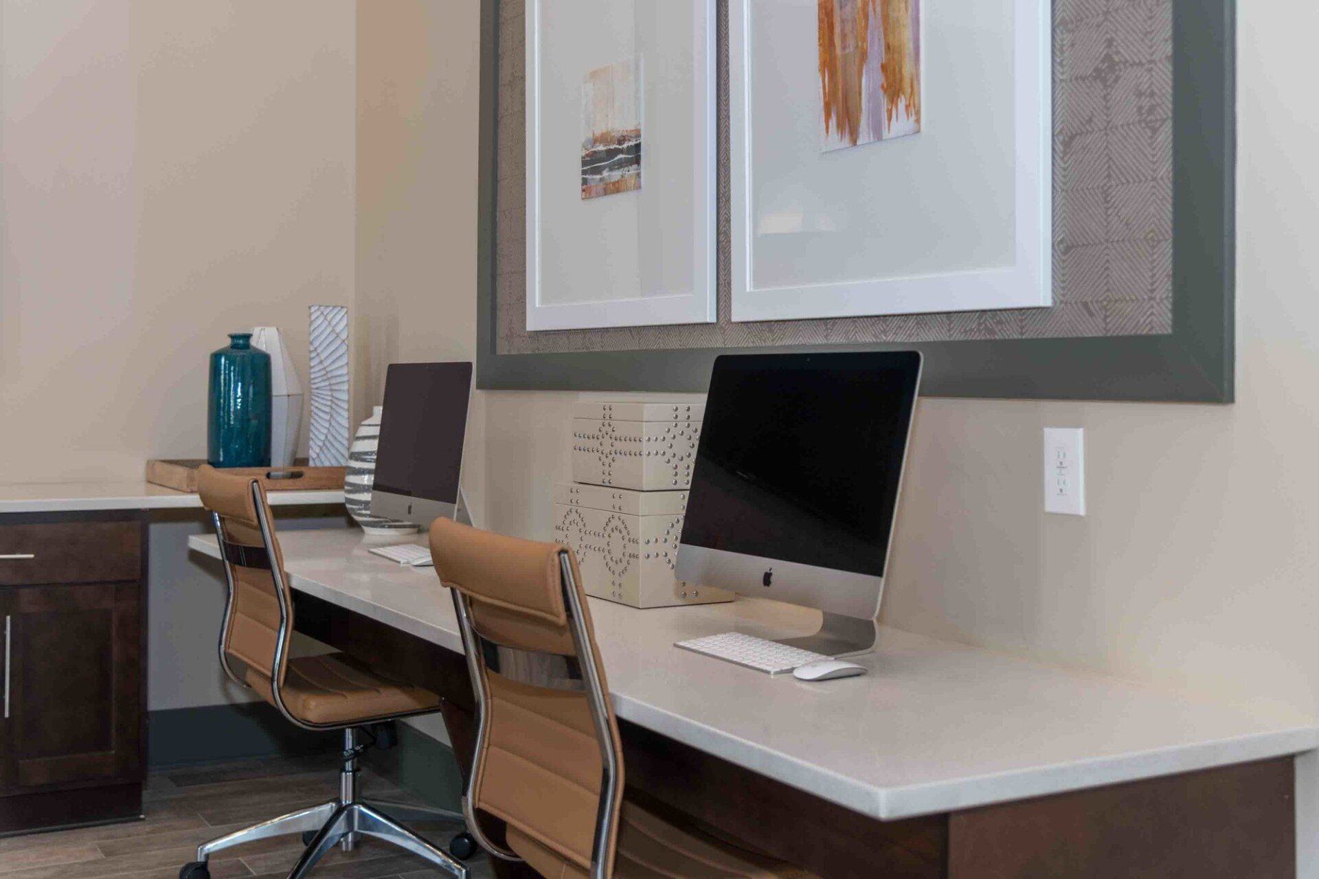 A modern home office with two workstations featuring computers, tan office chairs, and decorative items on a long desk at Silver Collection at Celebrate apartment community in Fredericksburg, VA.