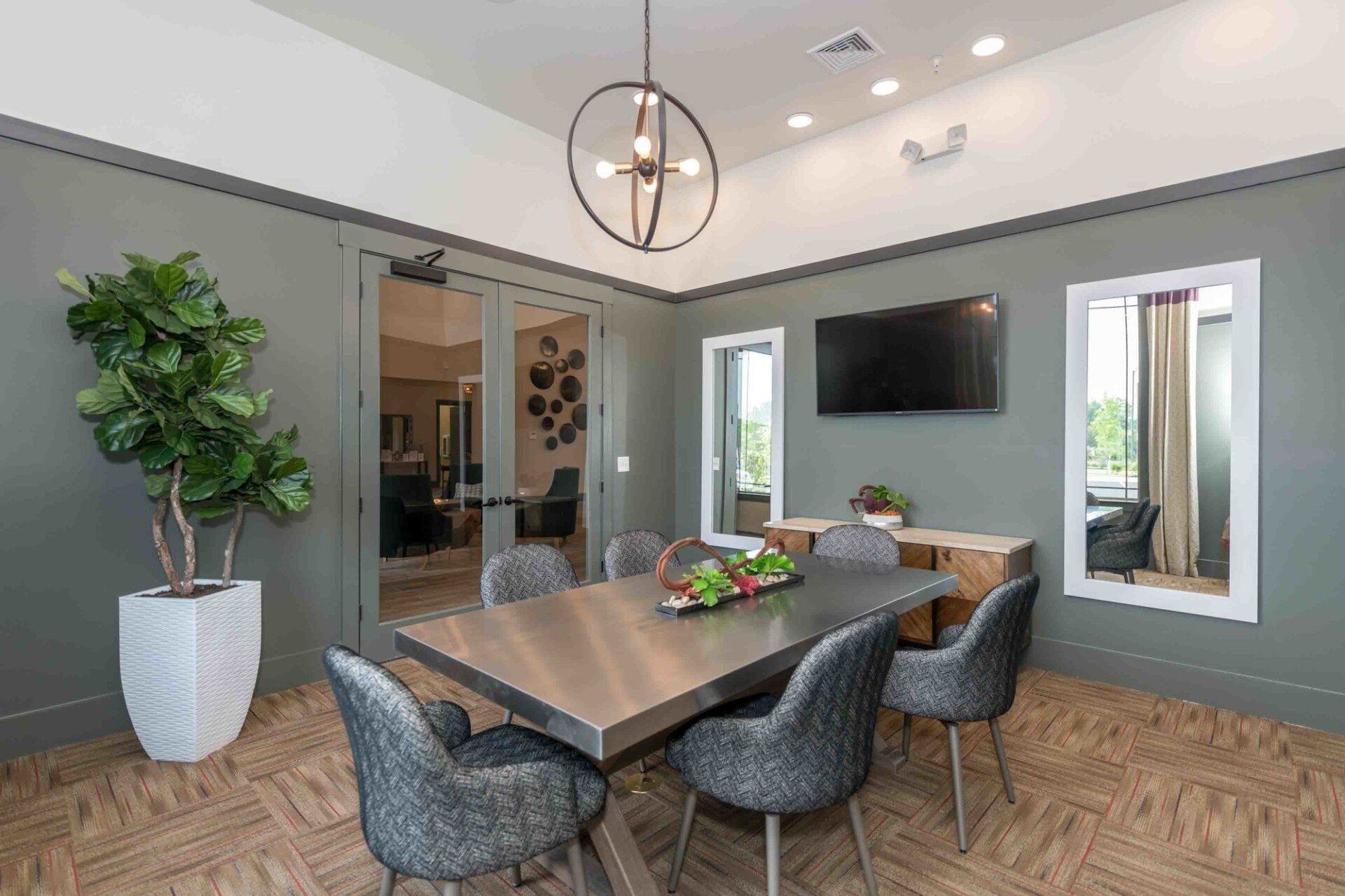 A modern conference room features a metal table with grey chairs, a green-painted wall, a TV, and a tall potted plant at Silver Collection at Celebrate apartment community in Fredericksburg, VA.