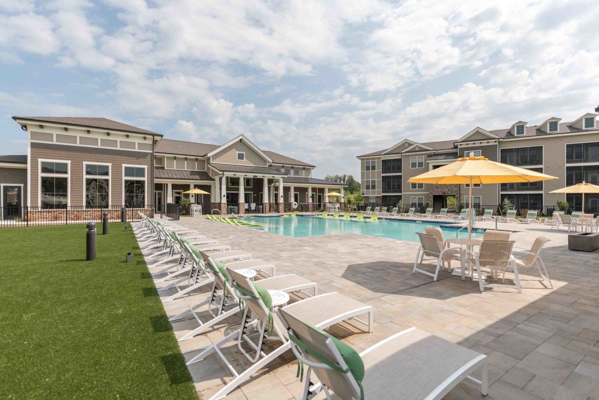 A resort-style pool area with lounge chairs, and umbrellas. Apartment buildings surround the pool under a partly cloudy sky.