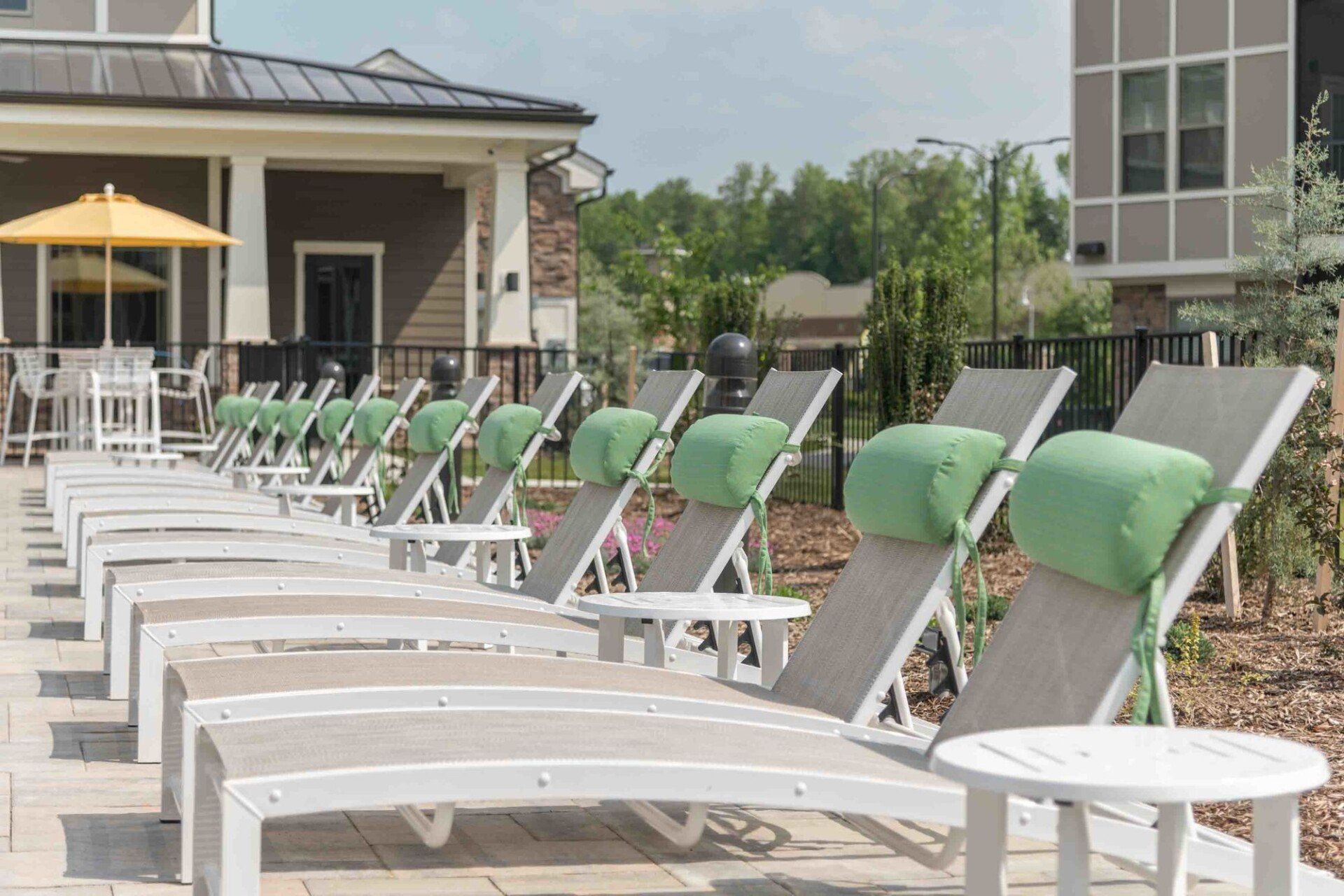 A row of lounge chairs with green headrest cushions lined up on a sunny patio next to a building and a yellow umbrella at Silver Collection at Celebrate apartment community in Fredericksburg, VA.