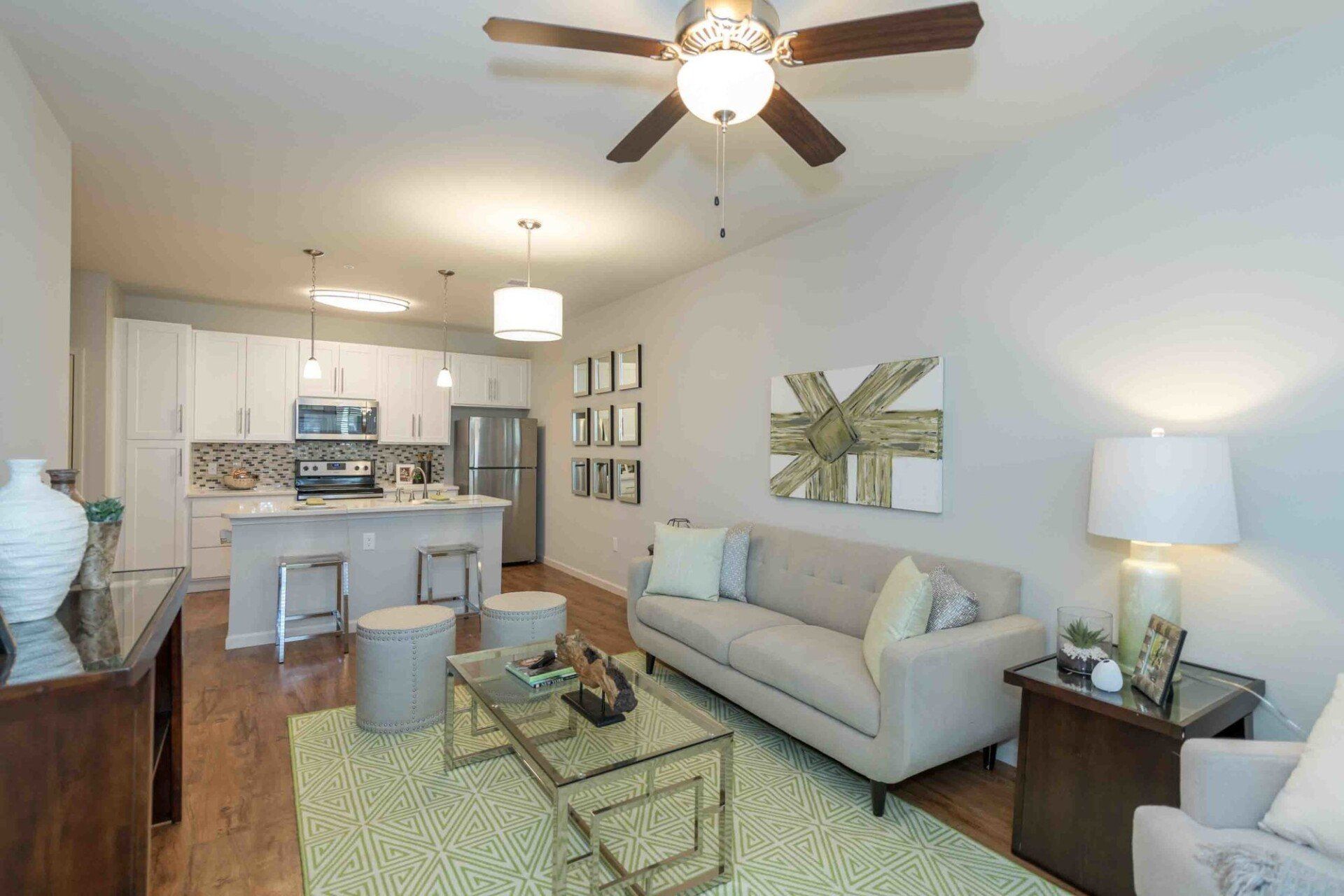 A modern open-plan living room with a beige sofa, glass coffee table, and an adjacent kitchen with a breakfast bar at Silver Collection at Celebrate apartment community in Fredericksburg, VA.