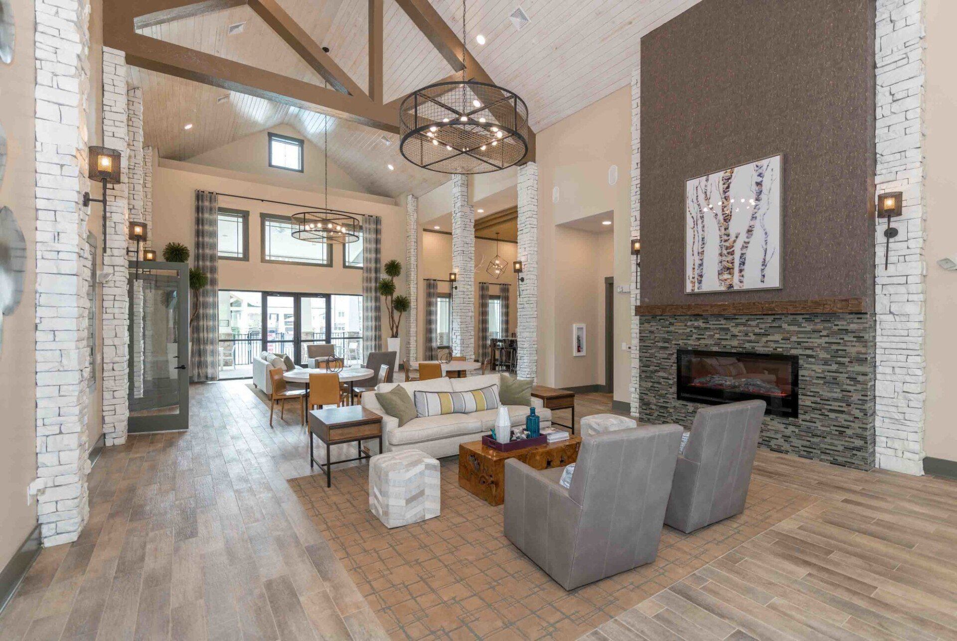 Lobby with fireplace, seating areas, and high ceilings. Features neutral tones, brick accents, and a modern chandelier.