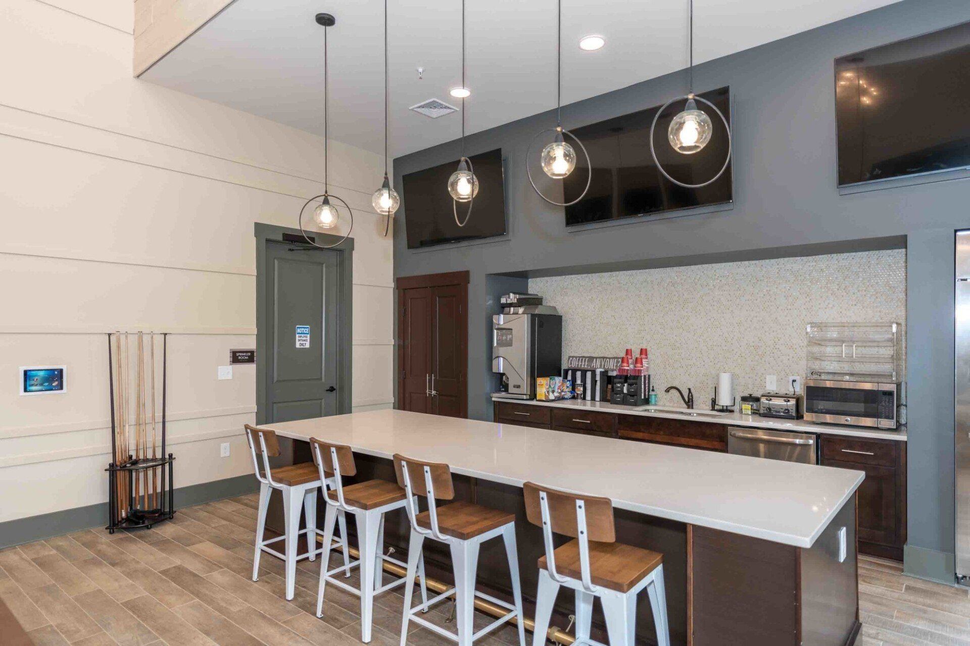 A communal kitchen area with a long counter, bar stools, pendant lighting, TVs mounted on a dark wall, and appliances at Silver Collection at Celebrate apartment community in Fredericksburg, VA.