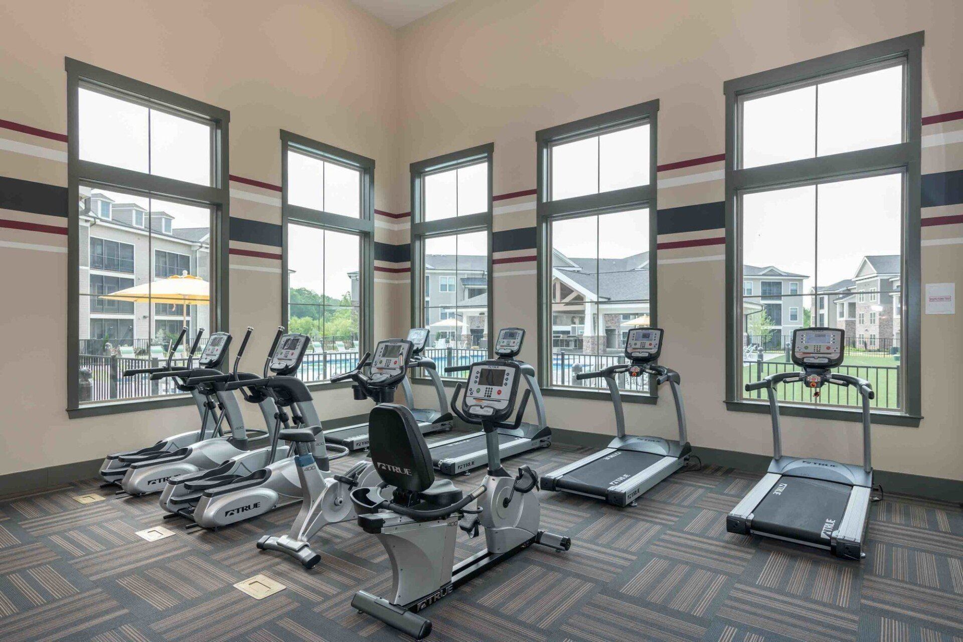 Gym with cardio equipment, including treadmills, and stationary bikes, facing large windows overlooking an outdoor area.