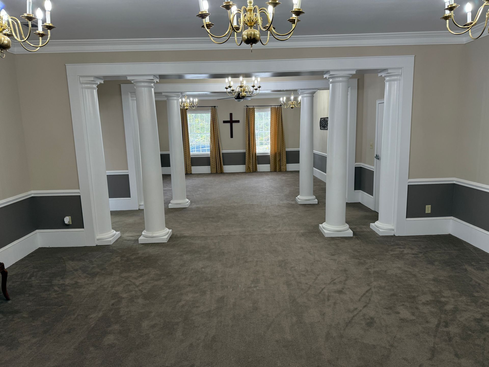 Empty church hall with white columns, gray carpet, chandeliers, and a cross at the far end.
