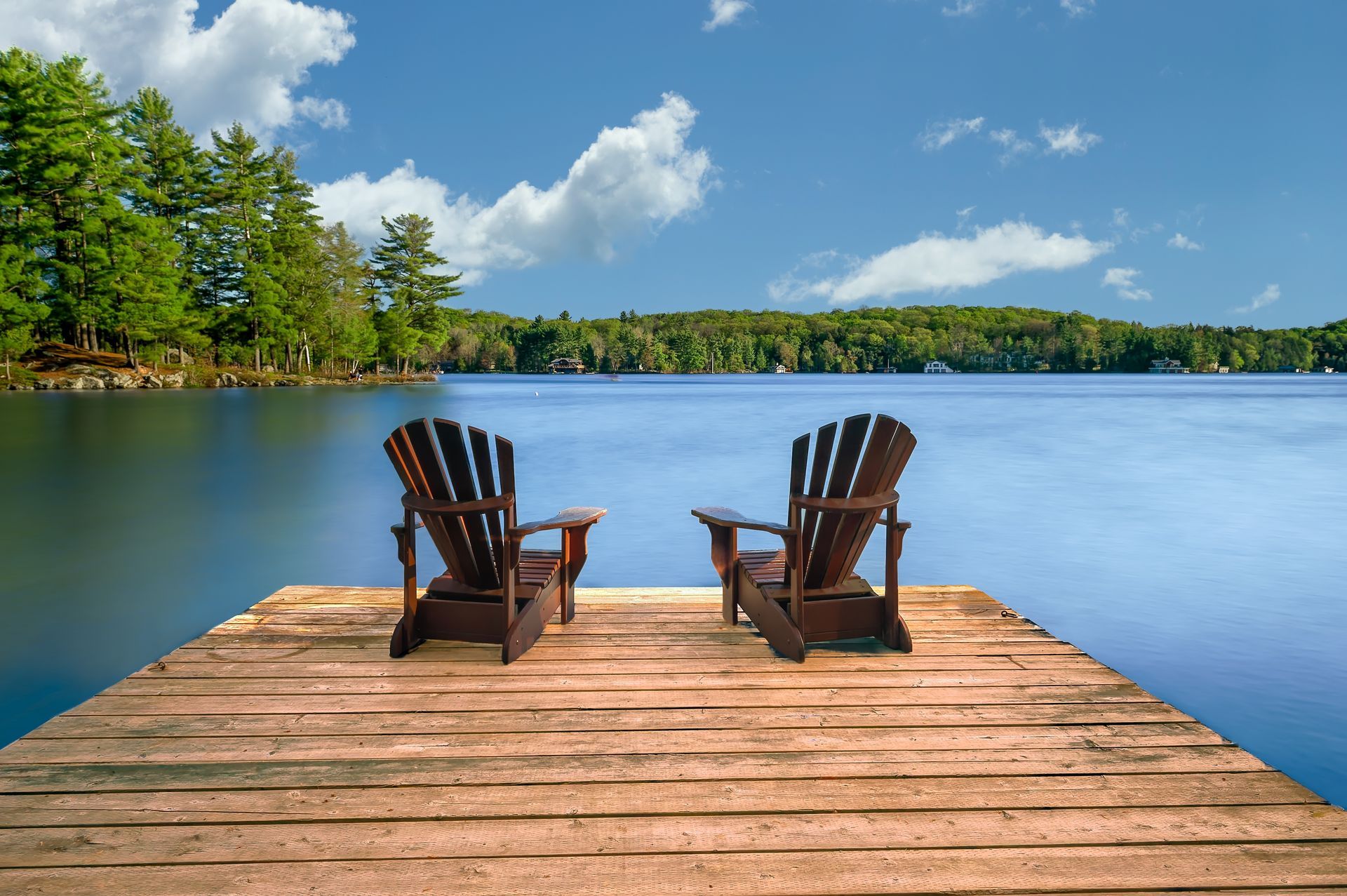 Two wooden chairs on a dock overlooking a calm blue lake and green trees under a sunny sky