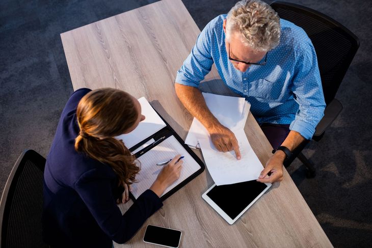 Two people review documents and a tablet at a conference table in an office.