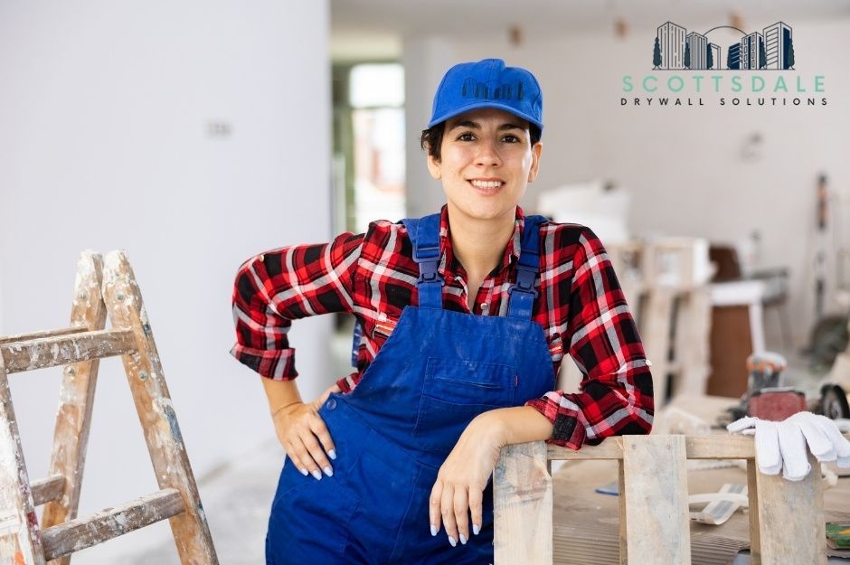 A drywall repair company worker, smiling, wearing a blue baseball cap, red plaid shirt, and blue overalls, leans on wooden sawhorses in a commercial construction site with a stepladder nearby and white walls in the background near W Flores Dr, and N 124th Ln, El Mirage, AZ.