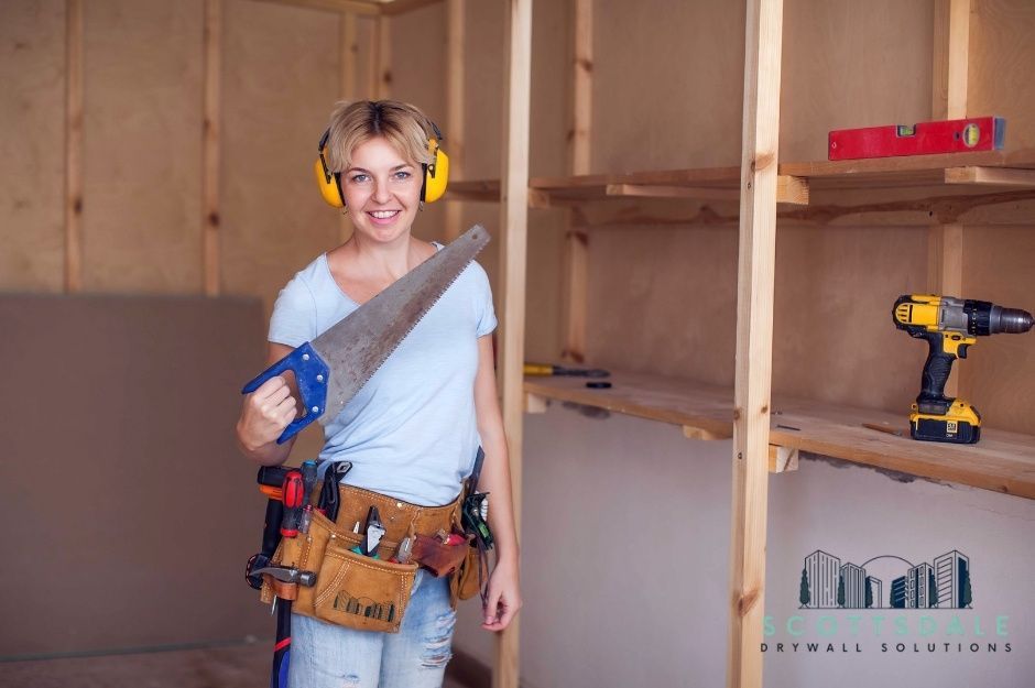 A drywall repair contractor wearing yellow ear protection and a tool belt holds a hand saw while working on a residential project with exposed wood framing. A cordless drill and a red level are visible on wooden shelves behind her. Located near N Crested Quail Run, and N Ocotillo Ridge Dr, Carefree, AZ.