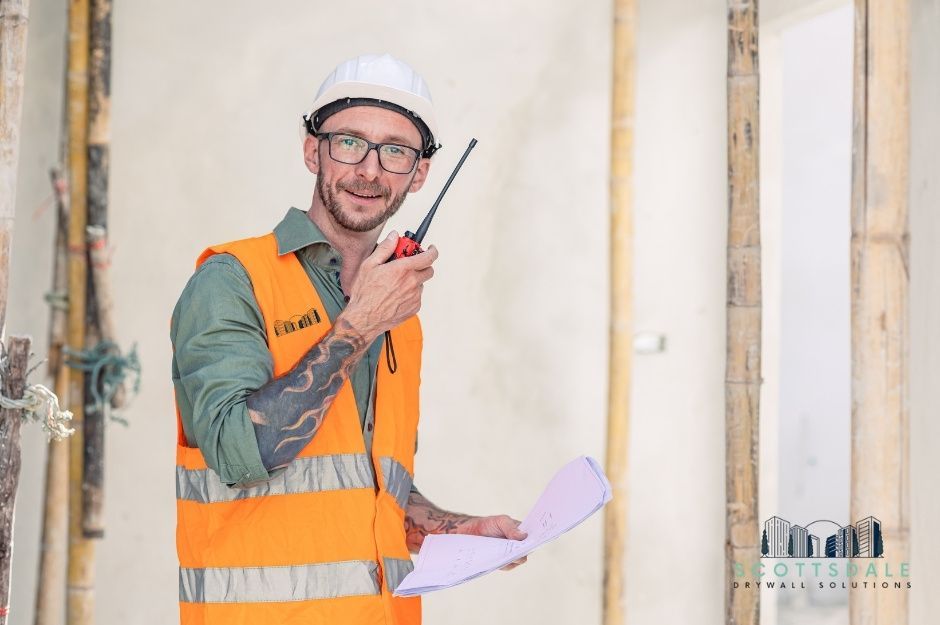 A drywall repair contractor in safety gear smiles while holding blueprints and a two-way radio at a residential construction site with exposed wooden framing and white drywall panels visible in the background near North 115th Avenue, and W Peoria Ave, Youngtown, AZ.