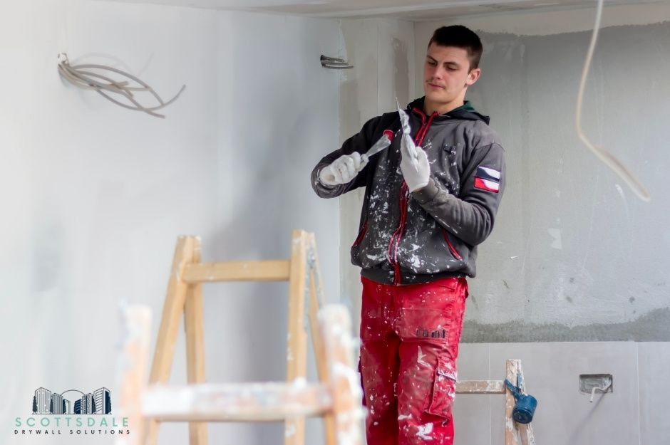A drywall repair handyman wearing red pants and a black jacket holds a putty knife while applying drywall compound to white walls. Paint splatters cover his work clothes as he repairs interior surfaces in a residential room near N 165th Dr, and W Woodlands Ave, Goodyear, AZ.